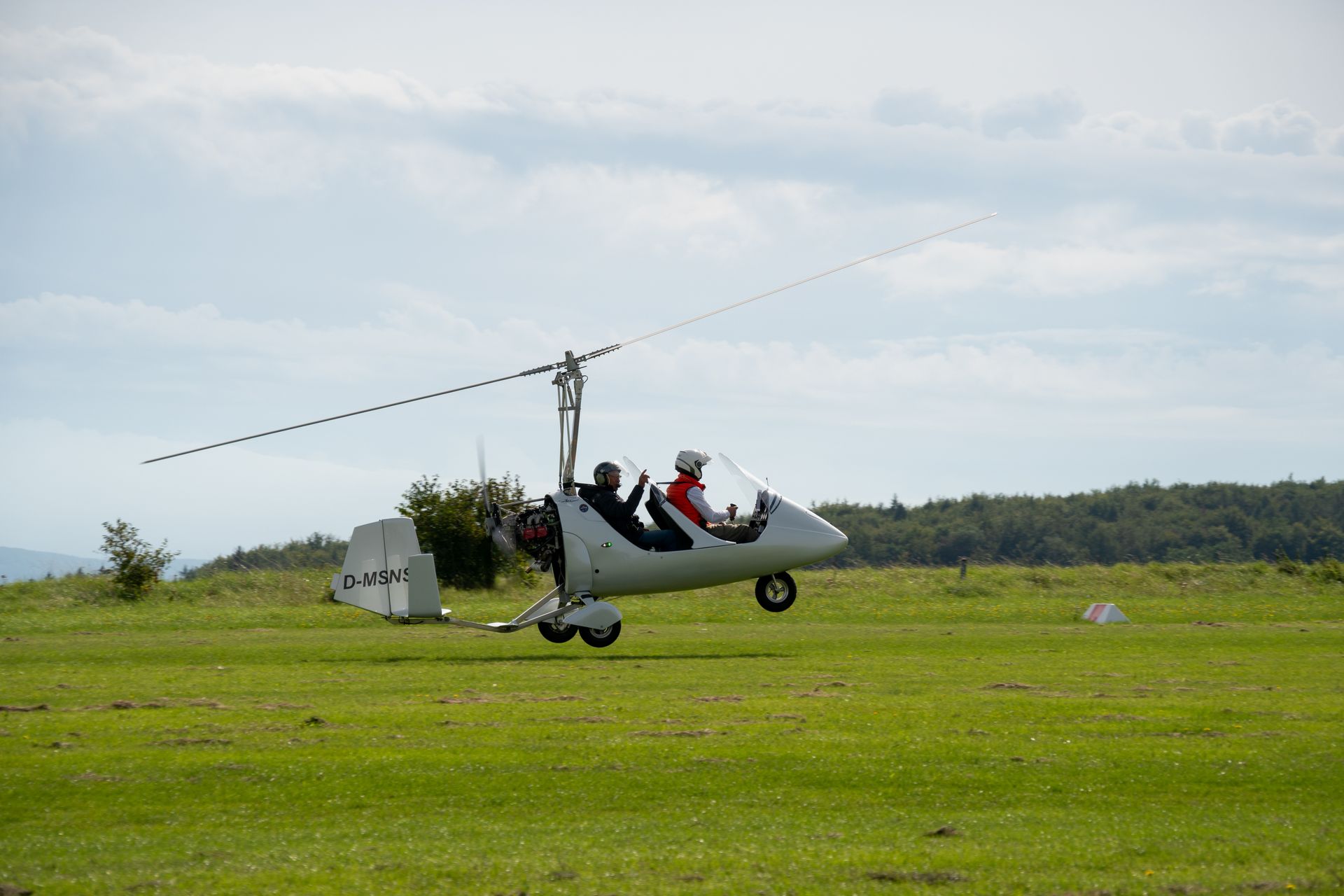 Ein Tragschrauber hebt unter bewölktem Himmel von einem Grasfeld ab. Pilot und Passagier im Cockpit.