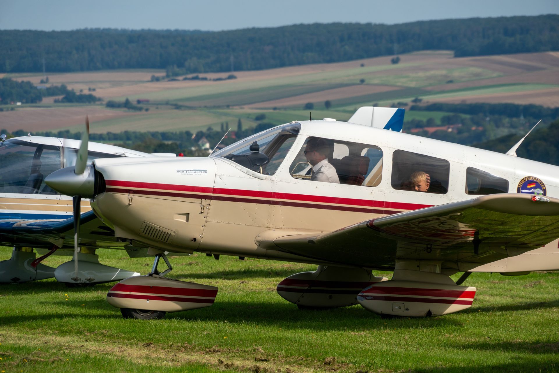 Weißes und rotes einmotoriges Flugzeug auf Gras. Pilot und Passagier im Cockpit, sanfte Hügel im Hintergrund.