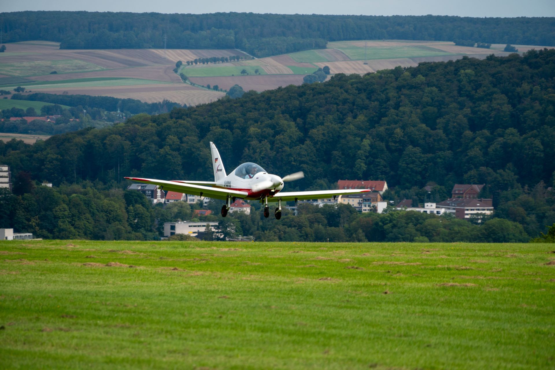 Ein kleines weißes Flugzeug landet auf einer grünen Grasfläche vor der Kulisse eines bewaldeten Hügels und Gebäuden.