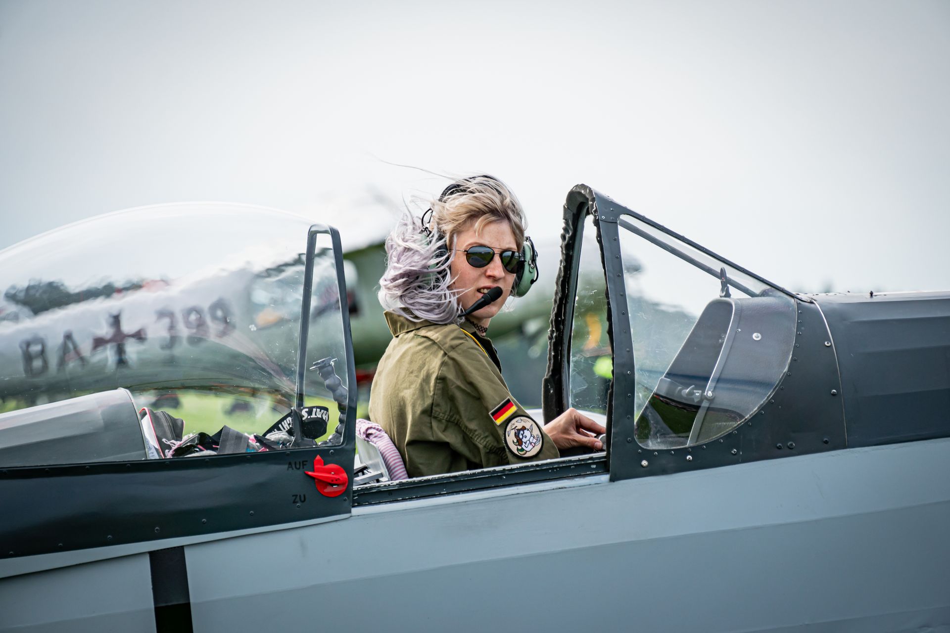 Pilot im offenen Cockpit, mit Headset und Sonnenbrille, mit einem Aufnäher der deutschen Flagge. Ein weiteres Flugzeug im Hintergrund.