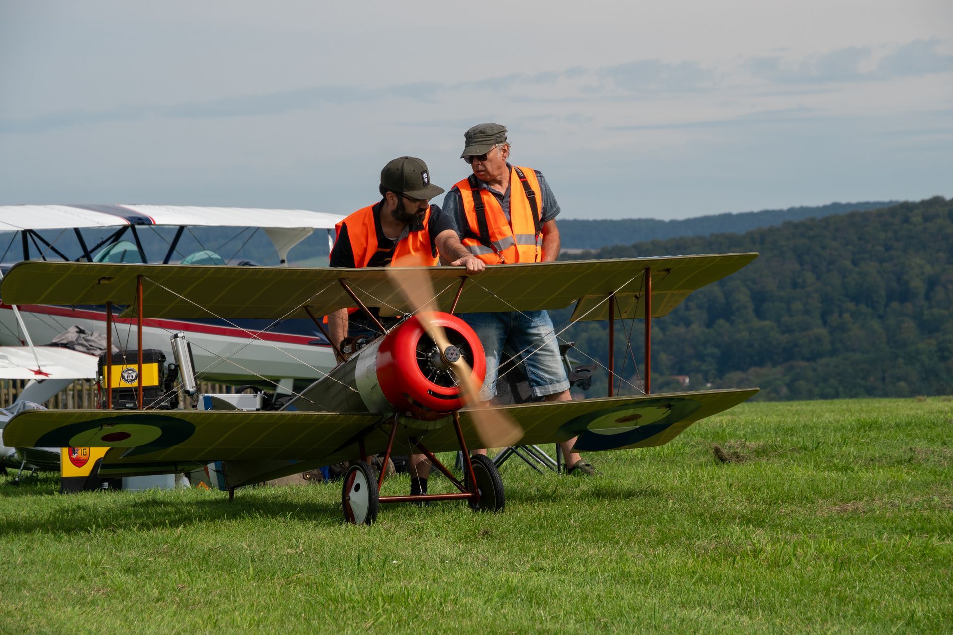 Zwei Männer begutachten ein hellbraunes Doppeldeckerflugzeug auf einem grasbewachsenen Feld; orangefarbene Westen, roter Propeller, grüne Hügel im Hintergrund.