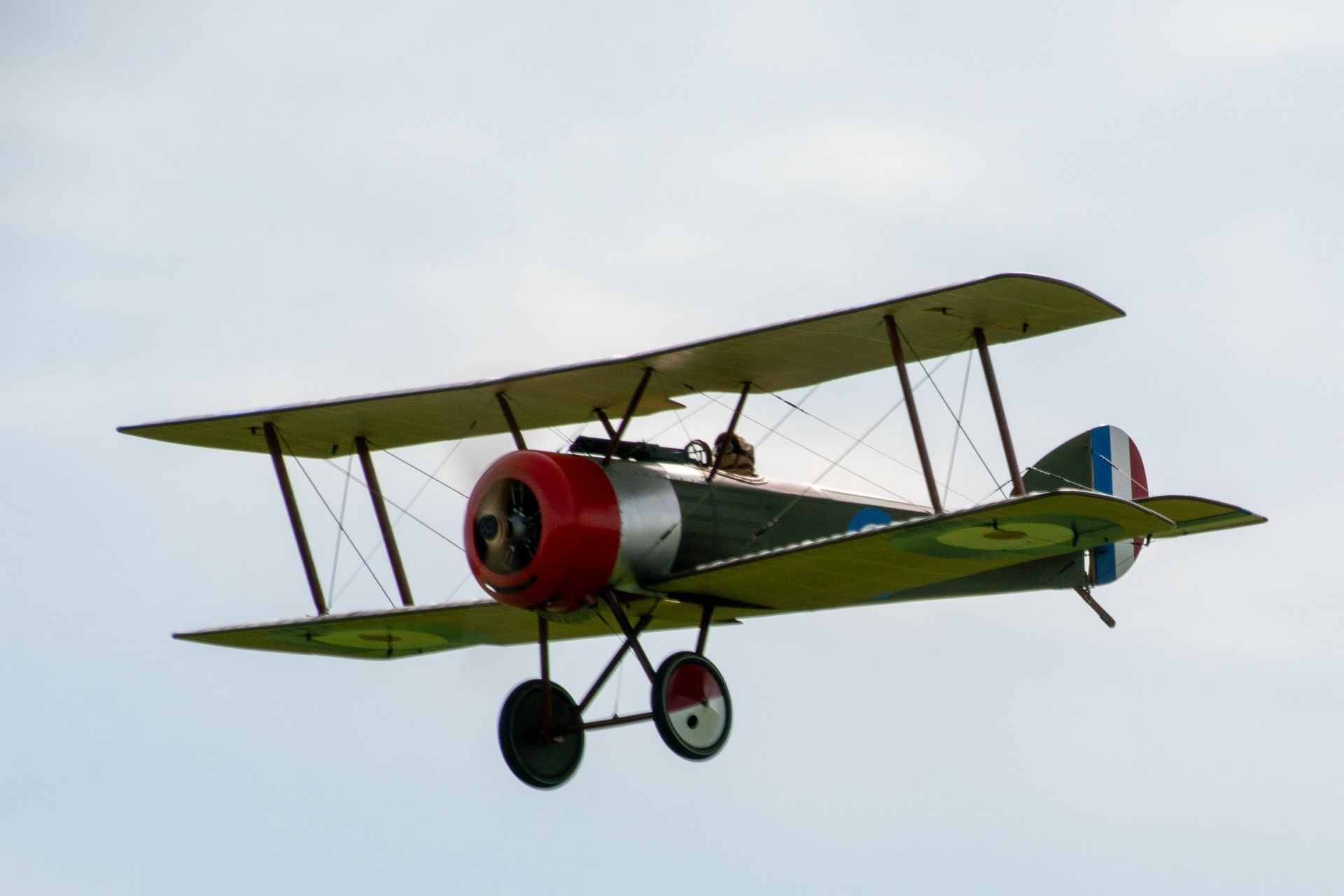 Doppeldecker im Flug mit roter Nase, silbernem Motor und grünen Tragflächen vor bewölktem Himmel.