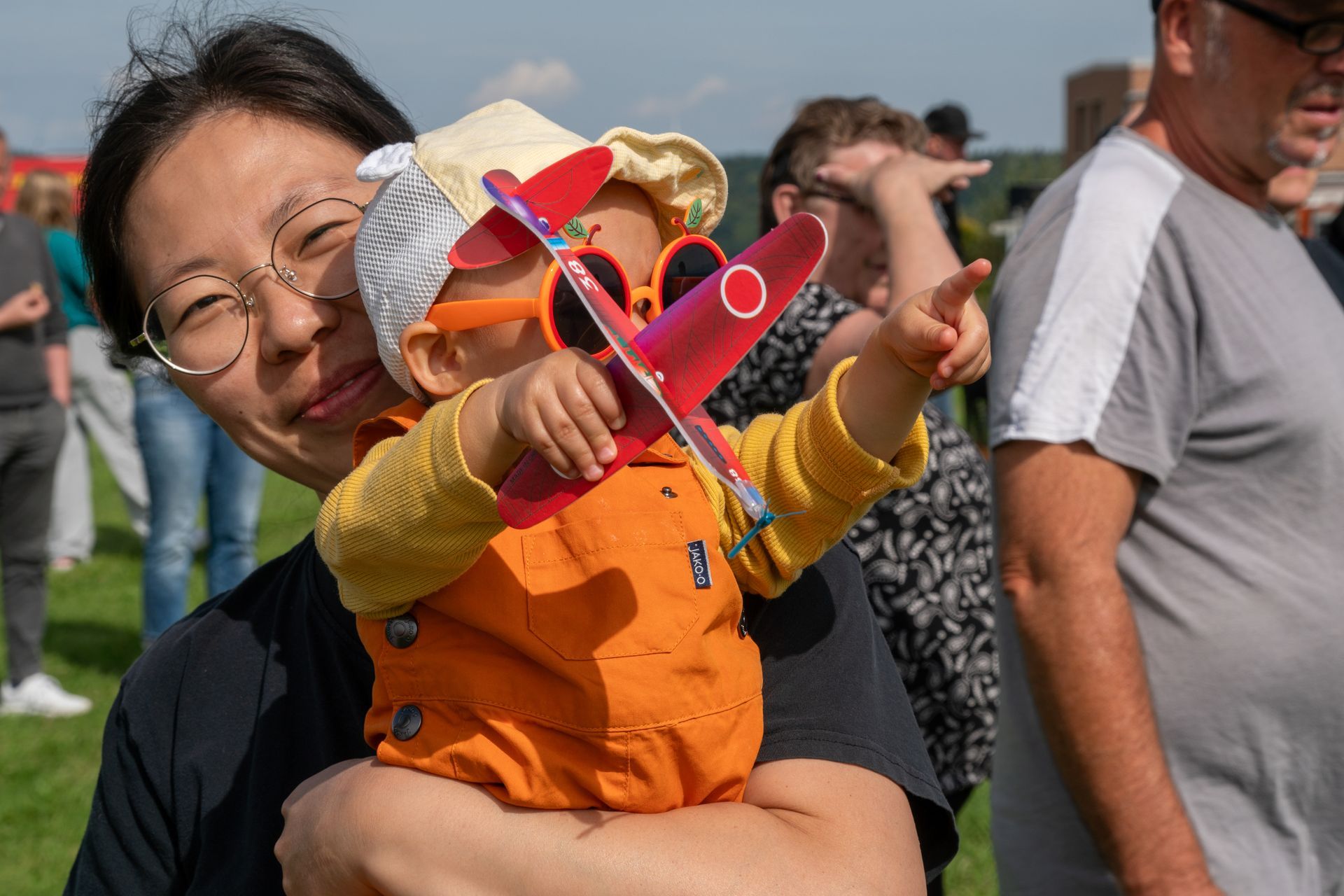 Frau hält ein Kind im Arm, das im Freien auf etwas zeigt; es spielt mit einem Spielzeugflugzeug.