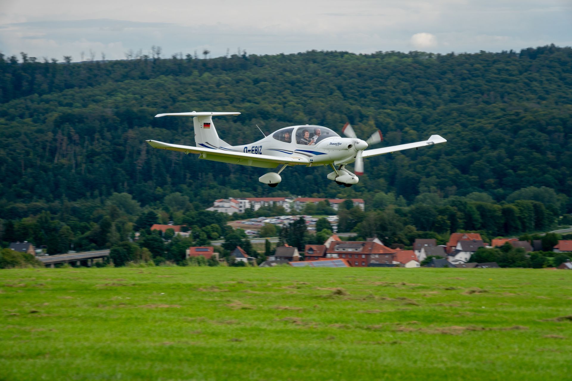 Ein weißes Flugzeug startet über einem grünen Feld; im Hintergrund sind ein Dorf und ein Wald zu sehen.