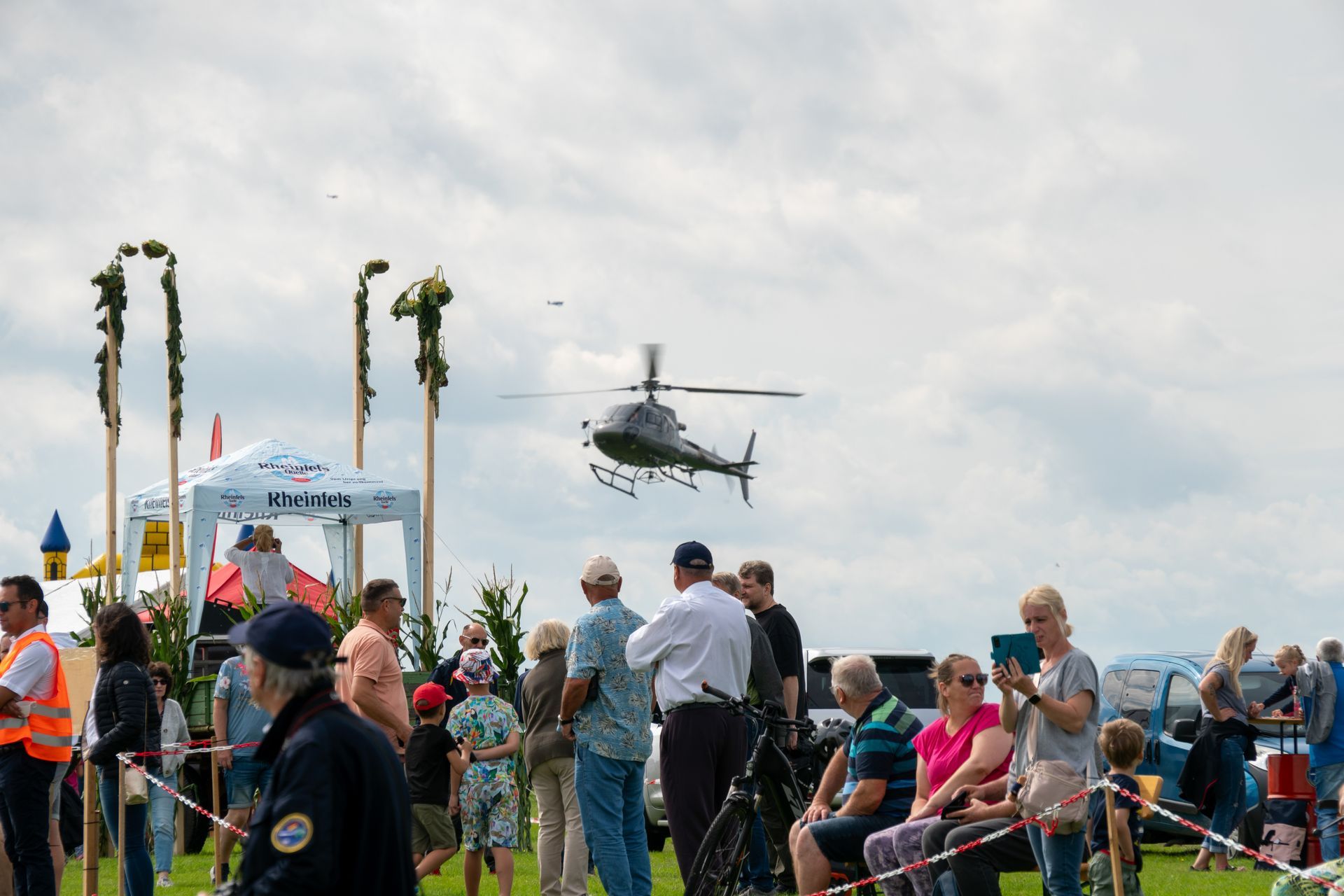 Menschen beobachten unter bewölktem Himmel einen Hubschrauber, der sich einem Ausstellungsbereich mit Zelten und Dekorationsstangen nähert.