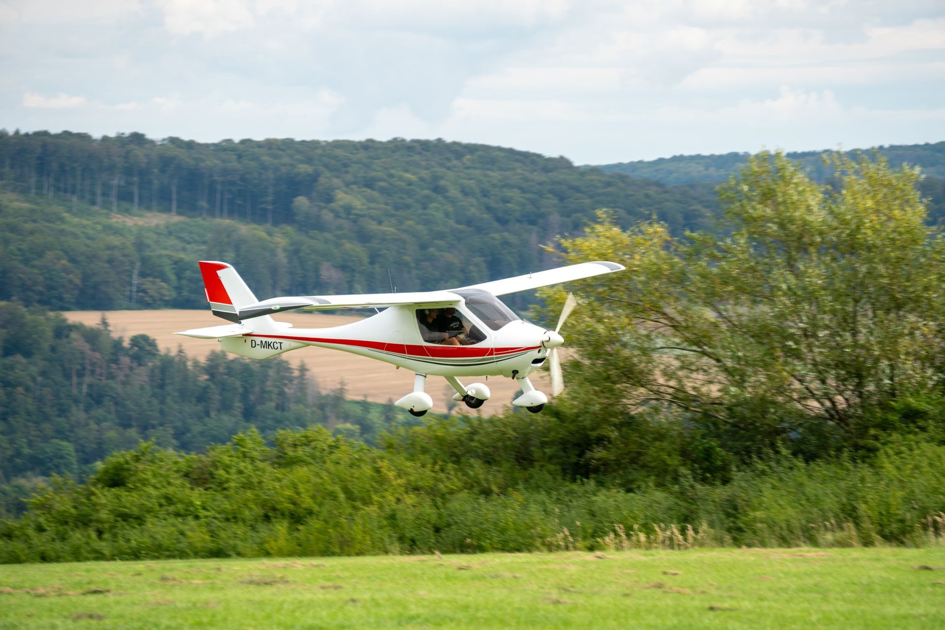 Ein weiß-rotes Flugzeug startet über einem grünen Feld, im Hintergrund sind Bäume und Hügel zu sehen.