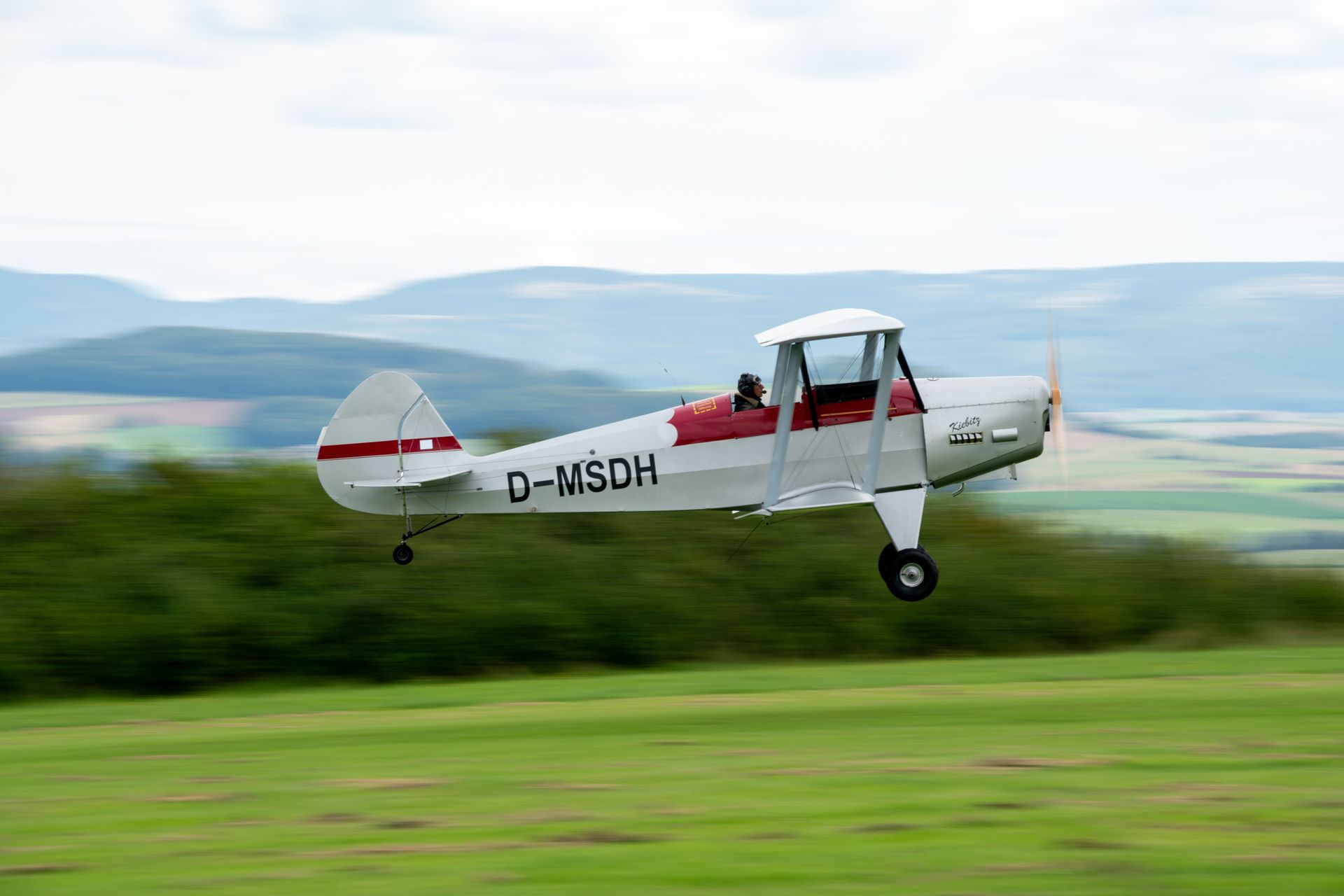 Ein weiß-rotes Doppeldeckerflugzeug startet über einem grünen Feld mit Bergen im Hintergrund.