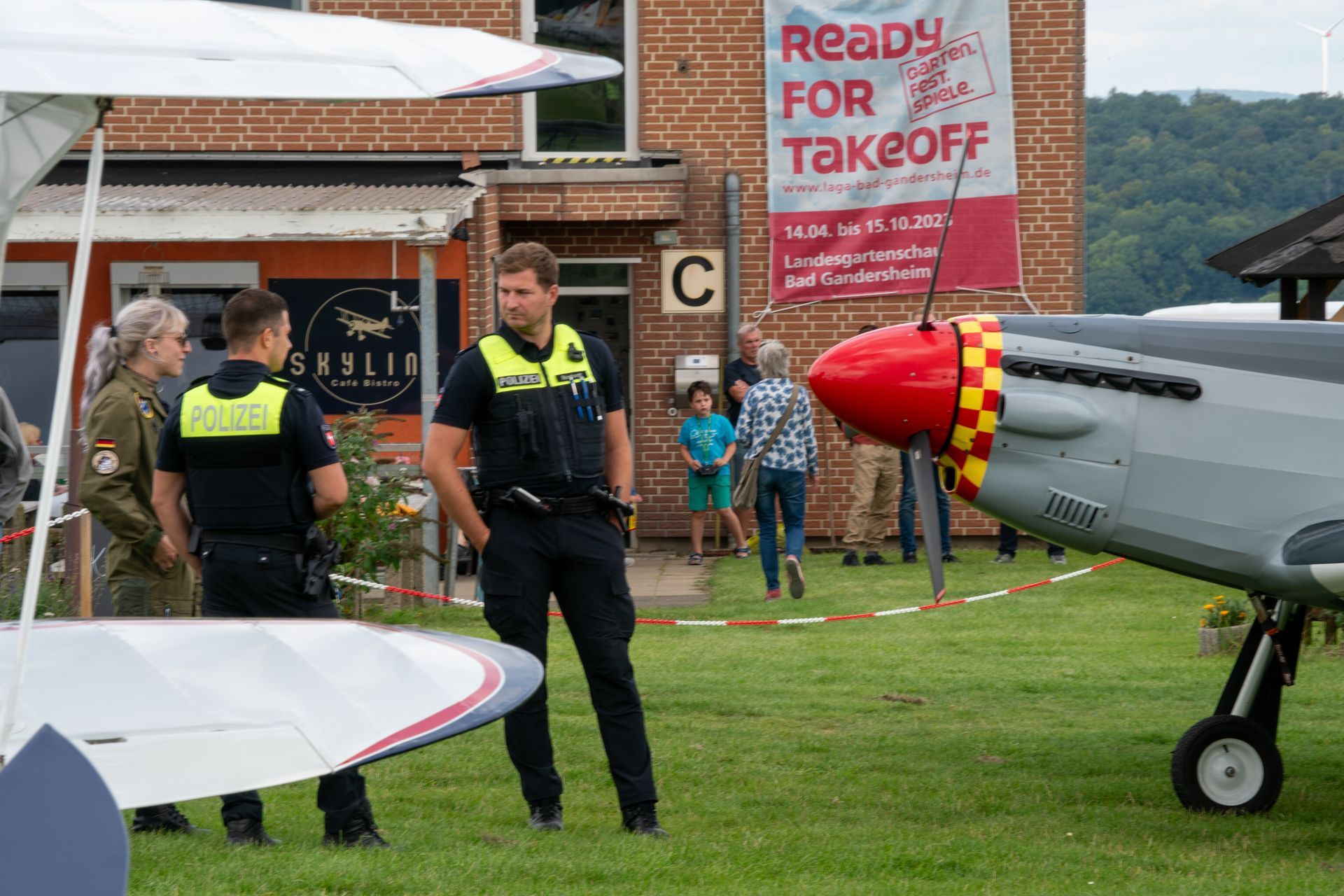 Zwei Polizisten und eine Person in Militäruniform stehen neben einem Flugzeug; auf einem Banner steht „Bereit zum Abheben“.