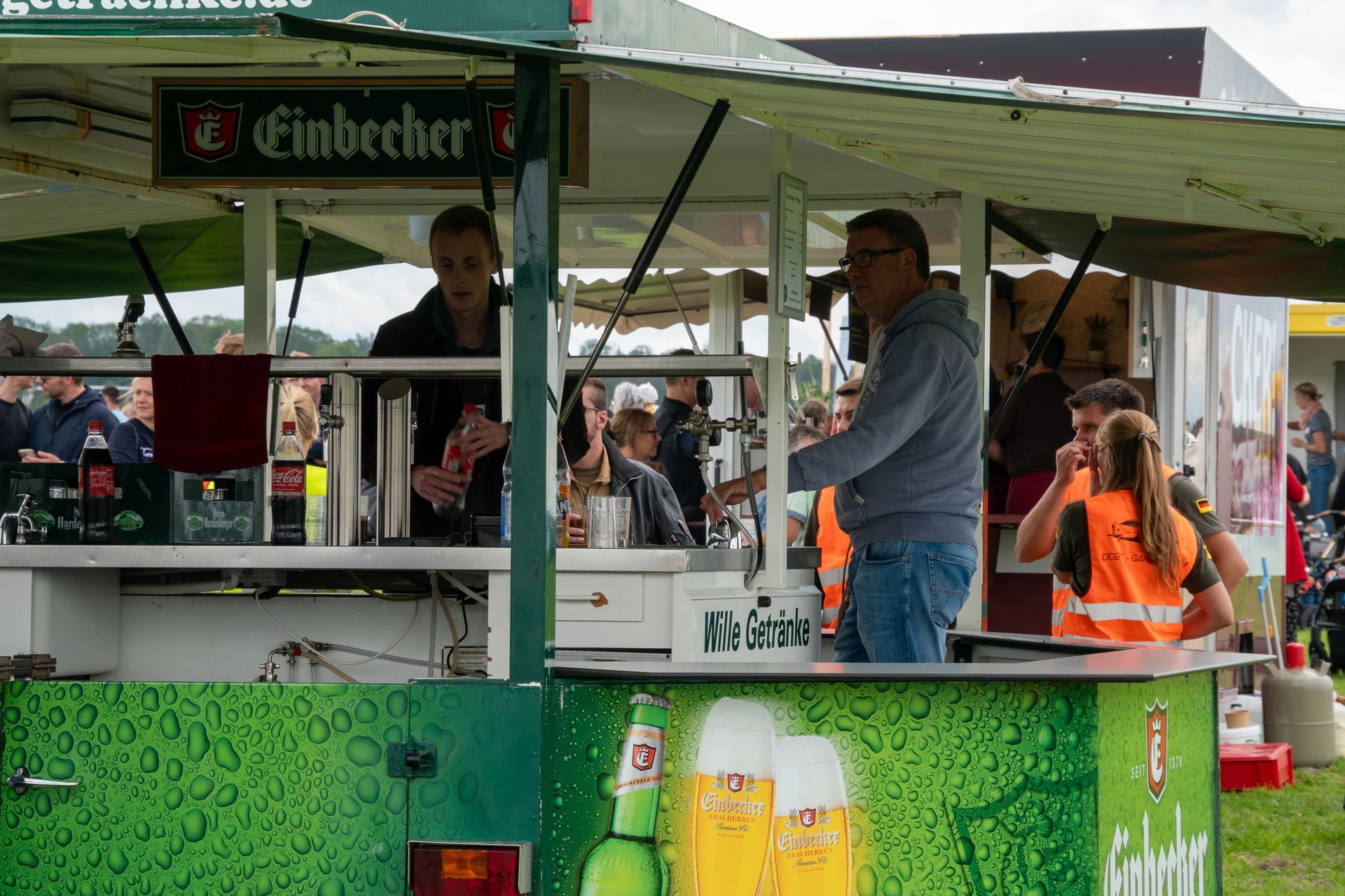 Ein Bierstand mit Leuten; ein Mann und eine Person in einer Weste sind zu sehen. Grüne, weiße und braune Farben.