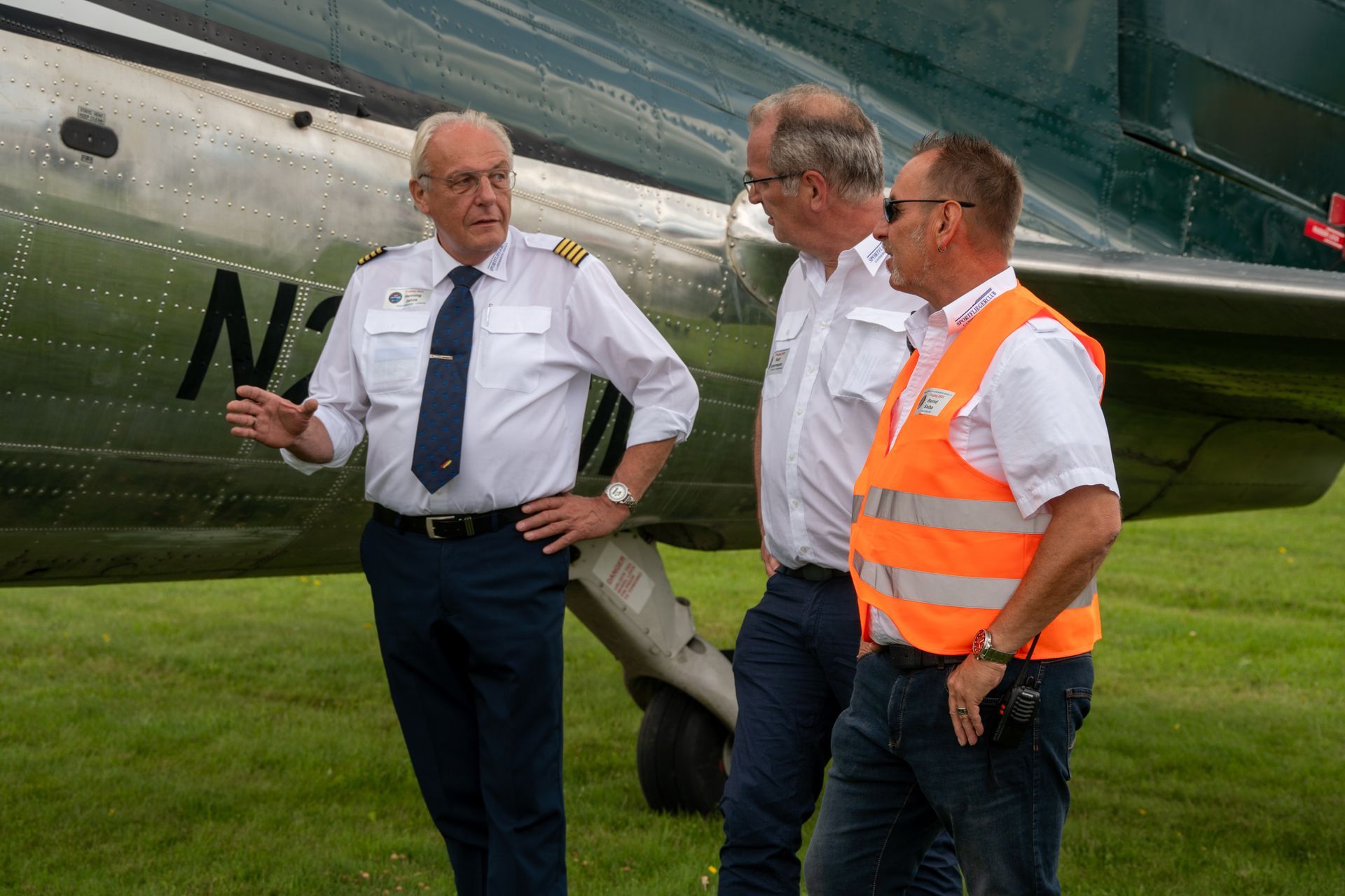Drei Männer unterhalten sich in der Nähe eines Flugzeugs auf einem grasbewachsenen Feld. Einer der Männer trägt eine Pilotenuniform.