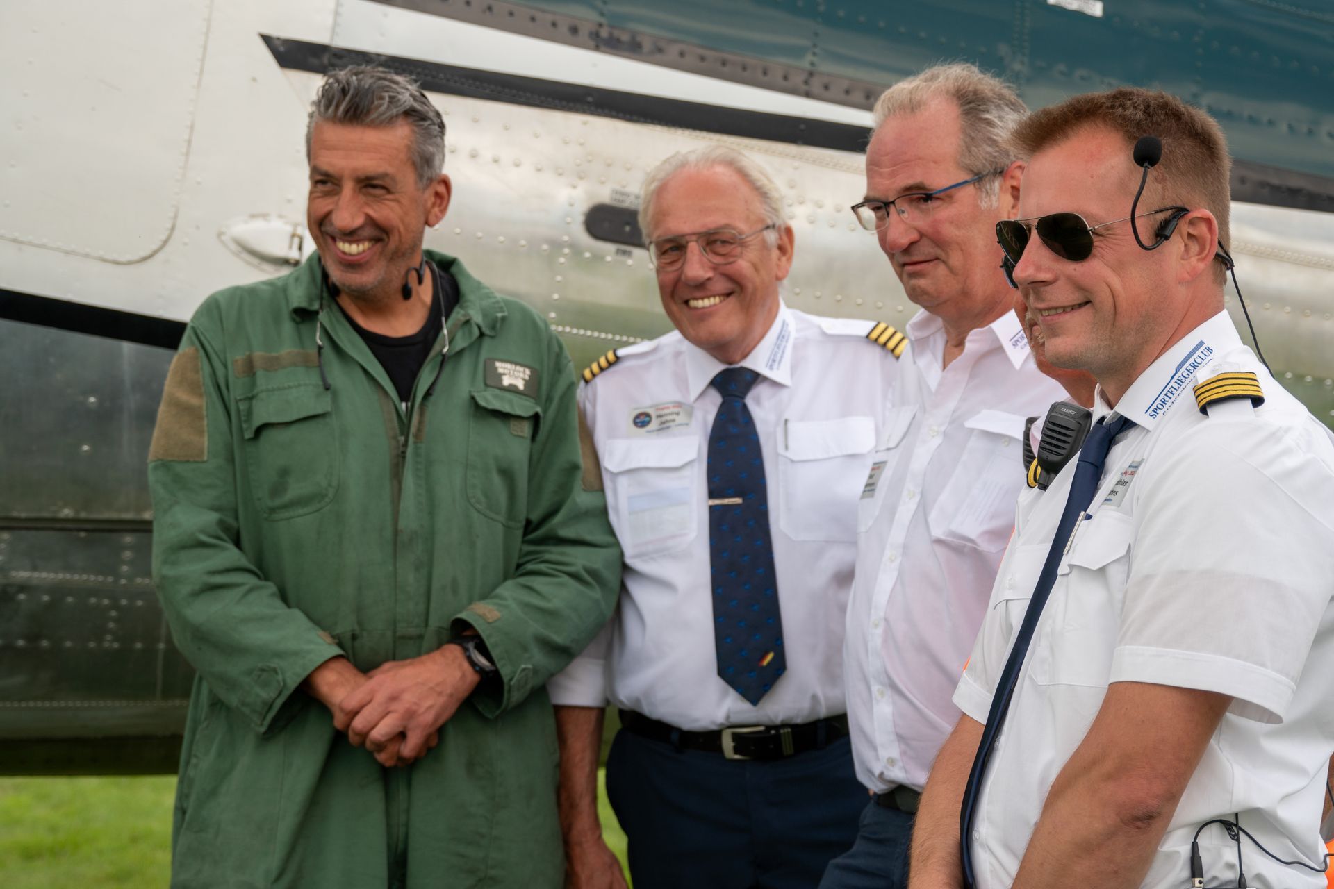 Vier Männer lächeln vor einem Flugzeug; einer trägt einen grünen Overall, zwei Pilotenuniformen, einer ein weißes Hemd.