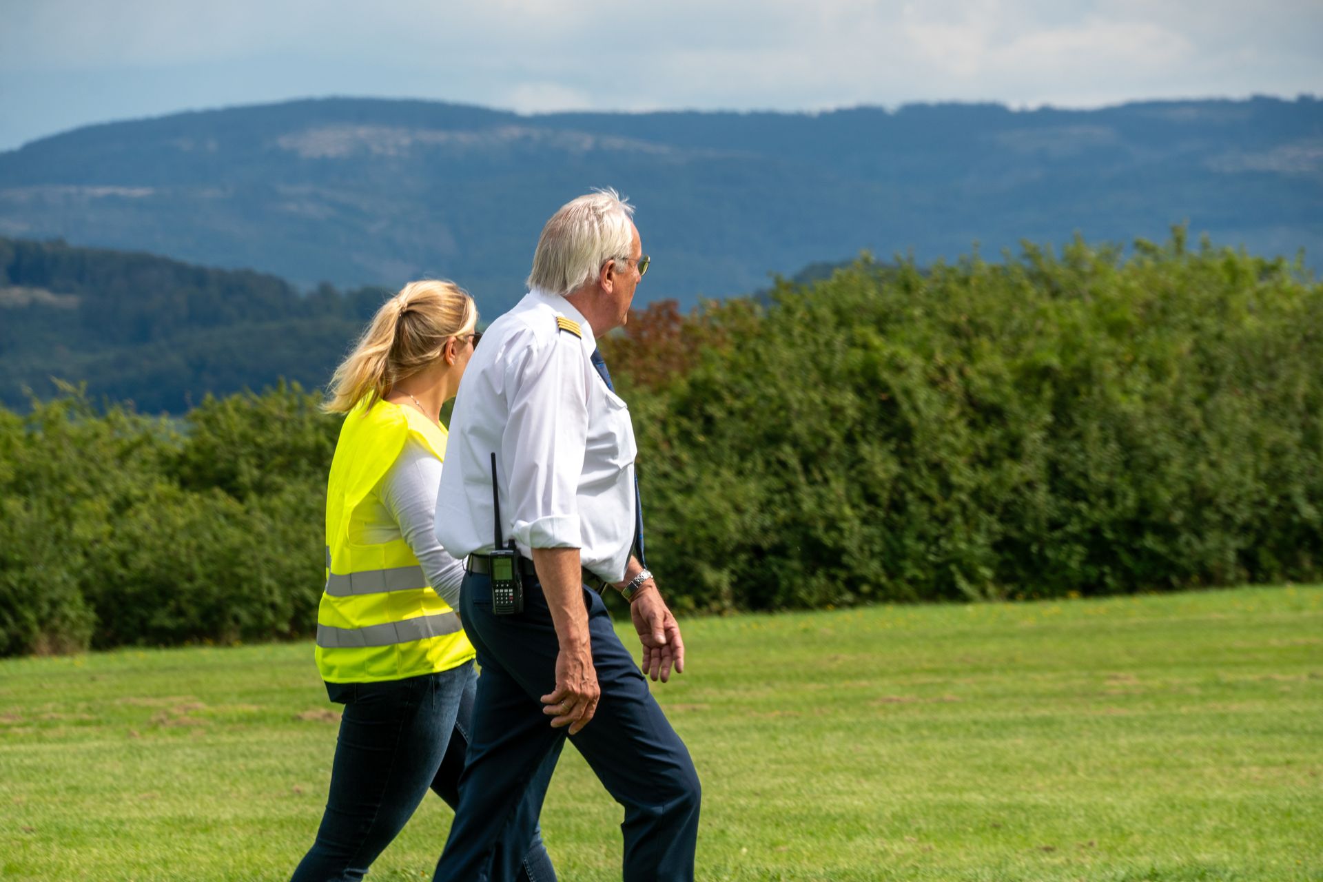 Zwei Personen gehen über eine Wiese. Eine trägt eine gelbe Warnweste, die andere ein weißes Hemd und eine dunkle Hose. Im Hintergrund sind Berge zu sehen.