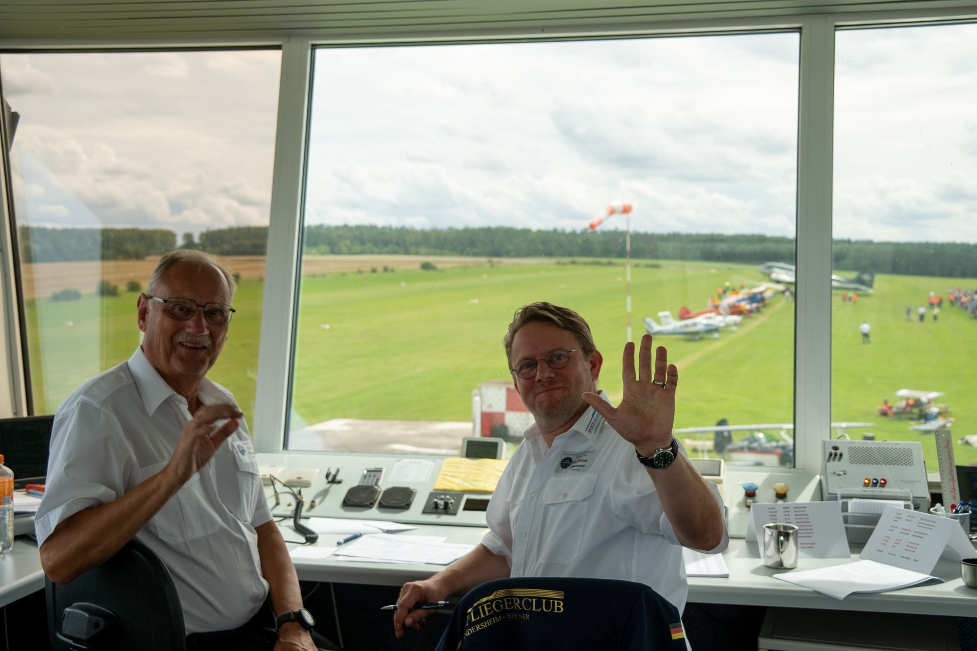 Zwei Männer in einem Fluglotsenturm mit Blick auf ein Flugfeld. Der eine winkt, der andere gestikuliert.
