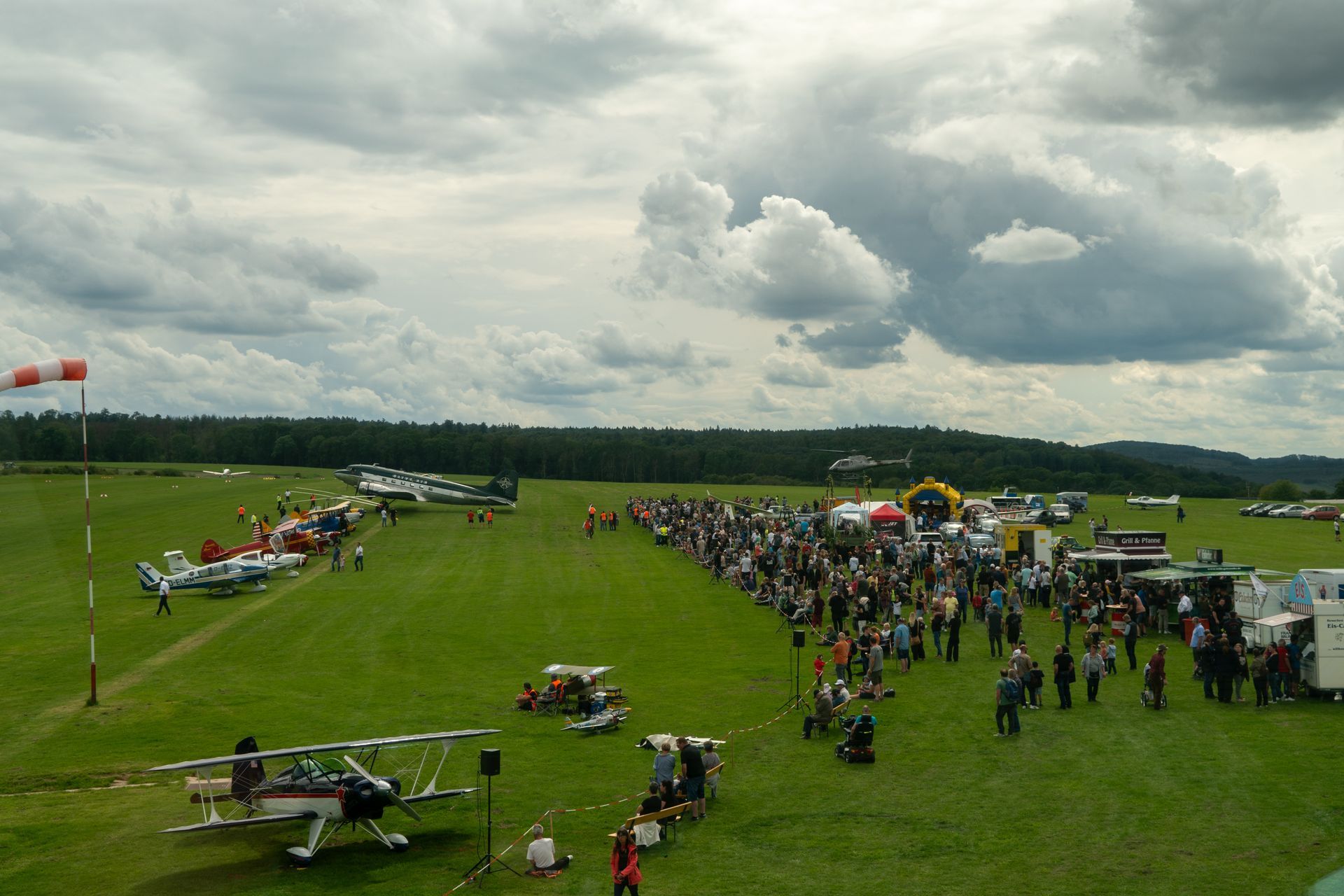 Zuschauermenge bei einer Flugschau auf einem grasbewachsenen Feld mit Flugzeugen; bewölkter Himmel darüber.