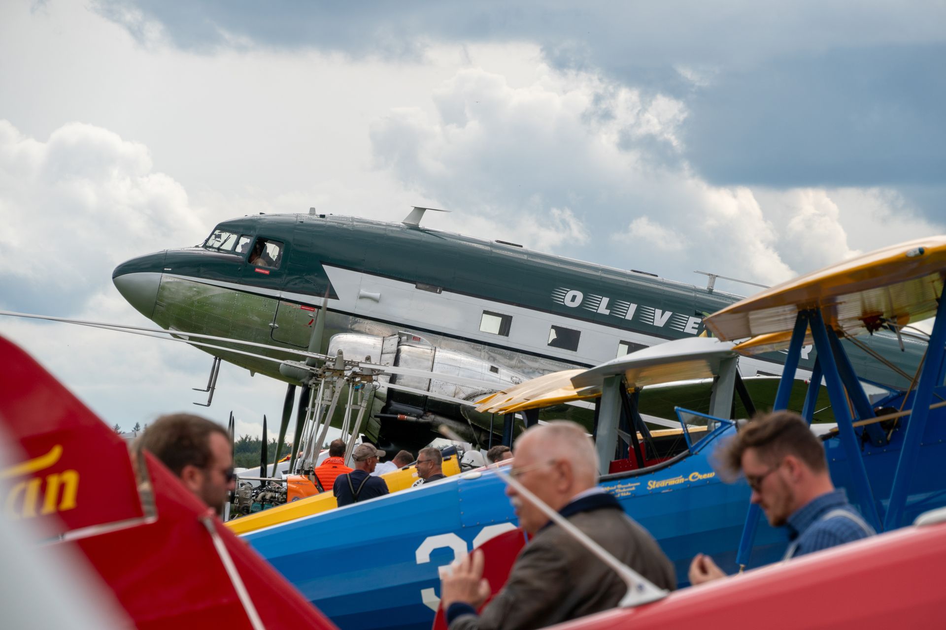 Ein älteres, grün-silbernes Flugzeug, geparkt mit anderen Flugzeugen, auf Augenhöhe zu sehen. Menschen stehen in der Nähe.