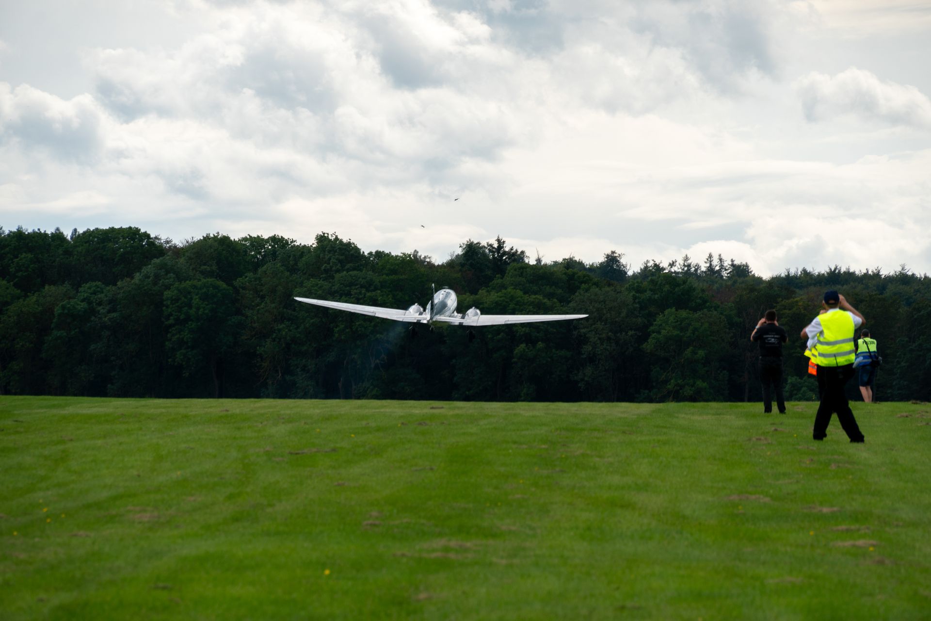Ein weißes Flugzeug landet auf einem grünen Grasfeld; im Hintergrund sind Bäume und auf der rechten Seite Menschen zu sehen.