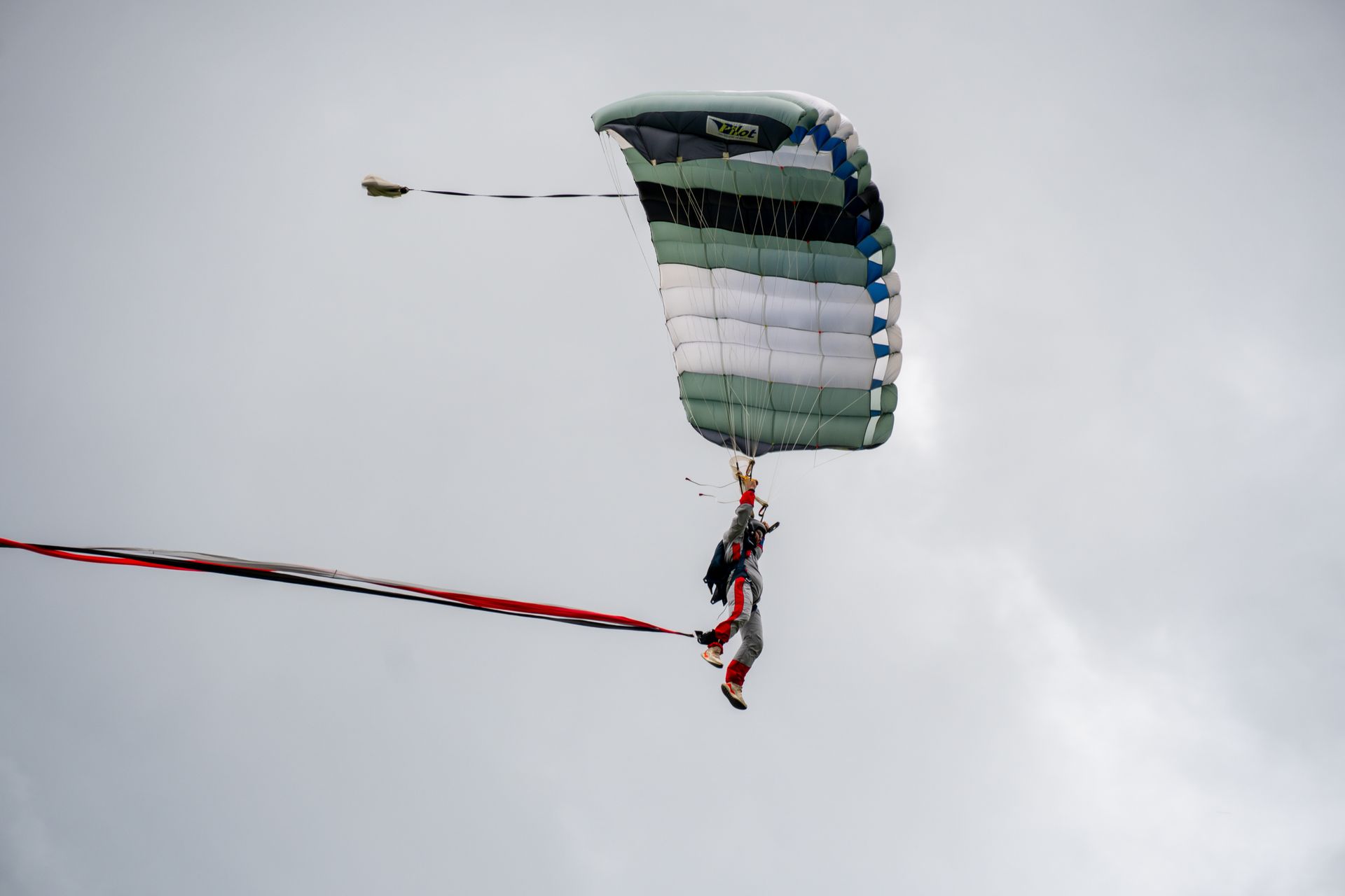 Ein Fallschirmspringer gleitet an einem grün-weißen Fallschirm herab, an dem eine rot-schwarze Schnur hängt. Bewölkter Himmel.
