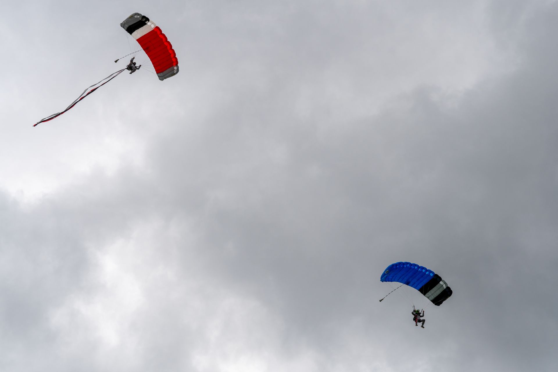 Zwei Fallschirmspringer steigen aus einem bewölkten Himmel herab. Der eine Fallschirm ist rot-schwarz, der andere blau-schwarz.