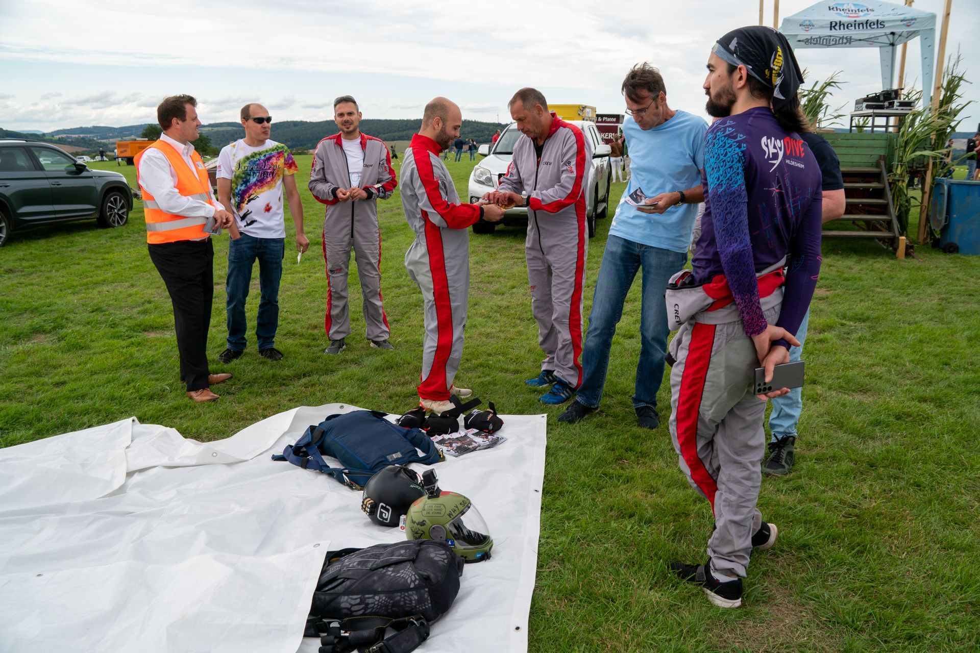 Eine Gruppe Fallschirmspringer in roten Anzügen bereitet ihre Ausrüstung auf einer Wiese vor. Ein Mann hält einen Helm, die anderen schauen zu.