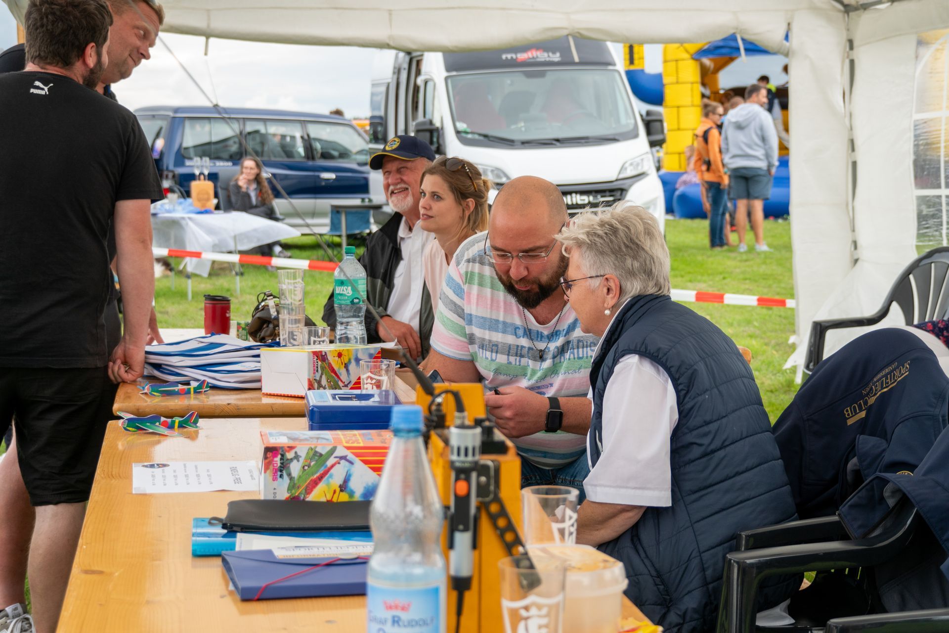 Menschen sitzen an einem Tisch unter einem Zelt und betrachten etwas. Im Hintergrund sind Flaschen, Kisten und ein Fahrzeug zu sehen.