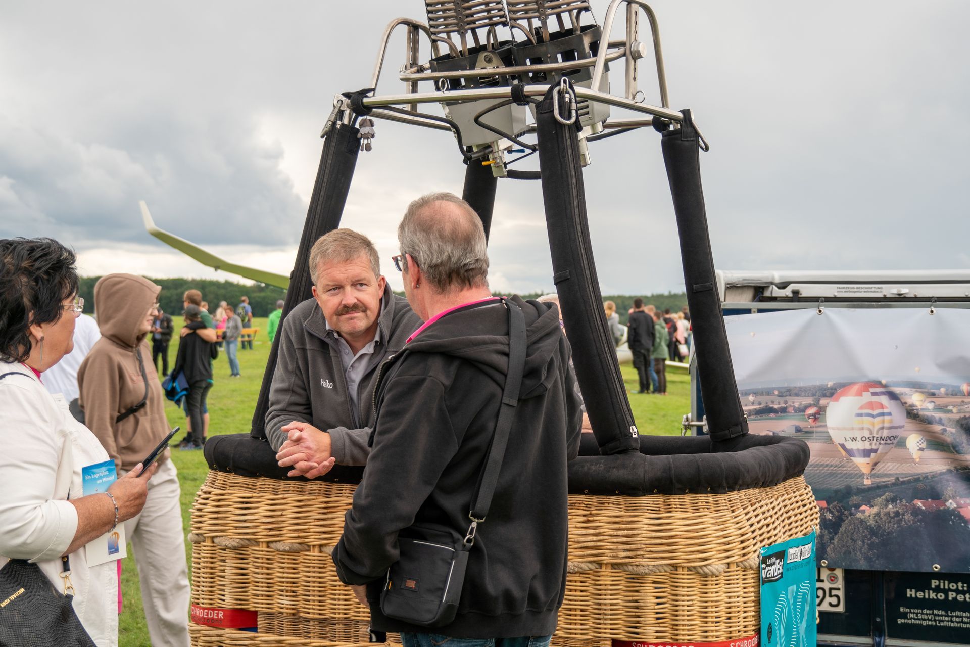 Menschen unterhalten sich neben dem Korb eines Heißluftballons; im Hintergrund sind ein grasbewachsenes Feld und ein bewölkter Himmel zu sehen.