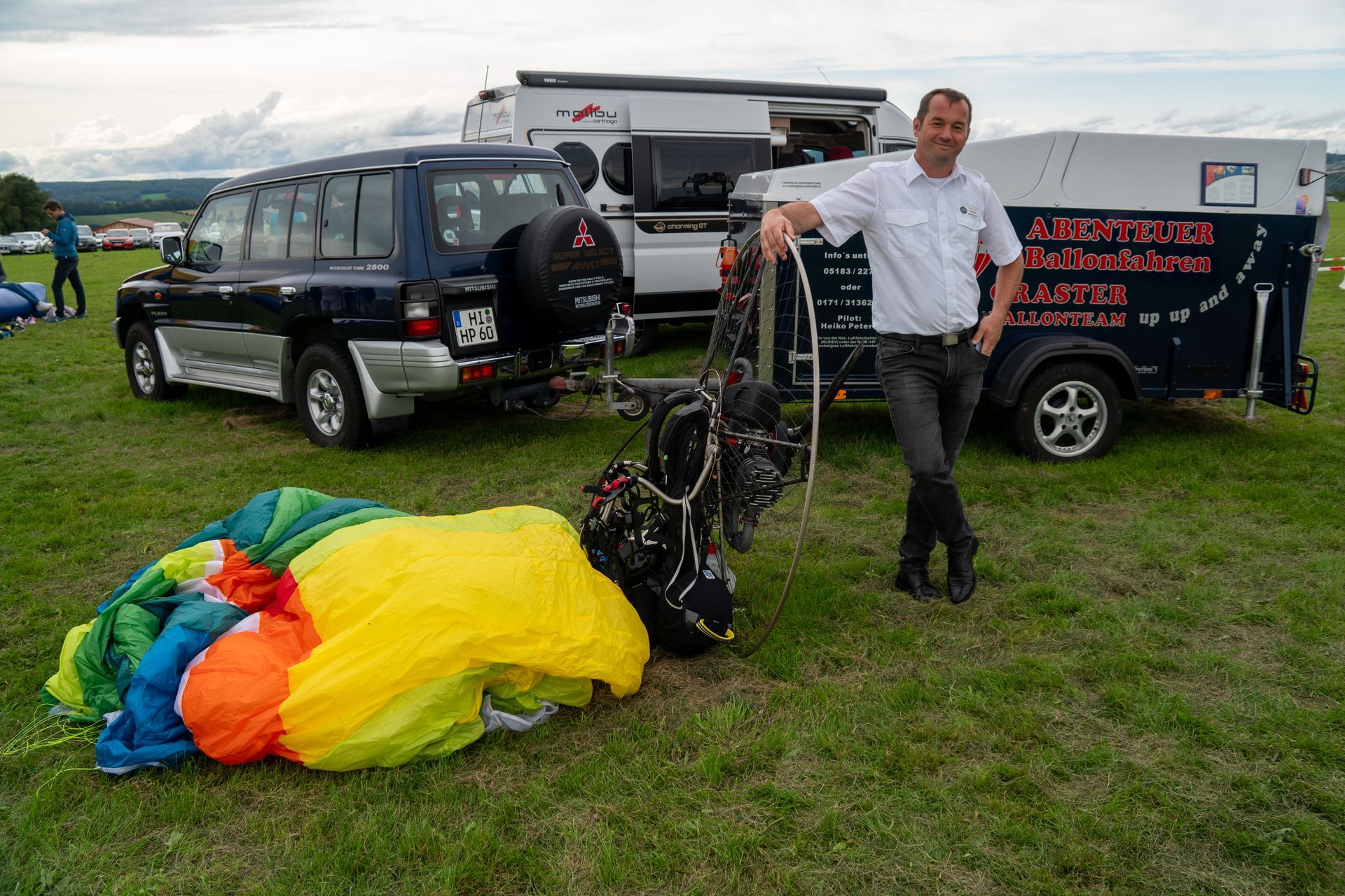Ein Mann mit Fallschirmausrüstung steht neben einem Anhänger und einem Fahrzeug auf einer Wiese. Ein bunter Fallschirm liegt auf dem Boden.