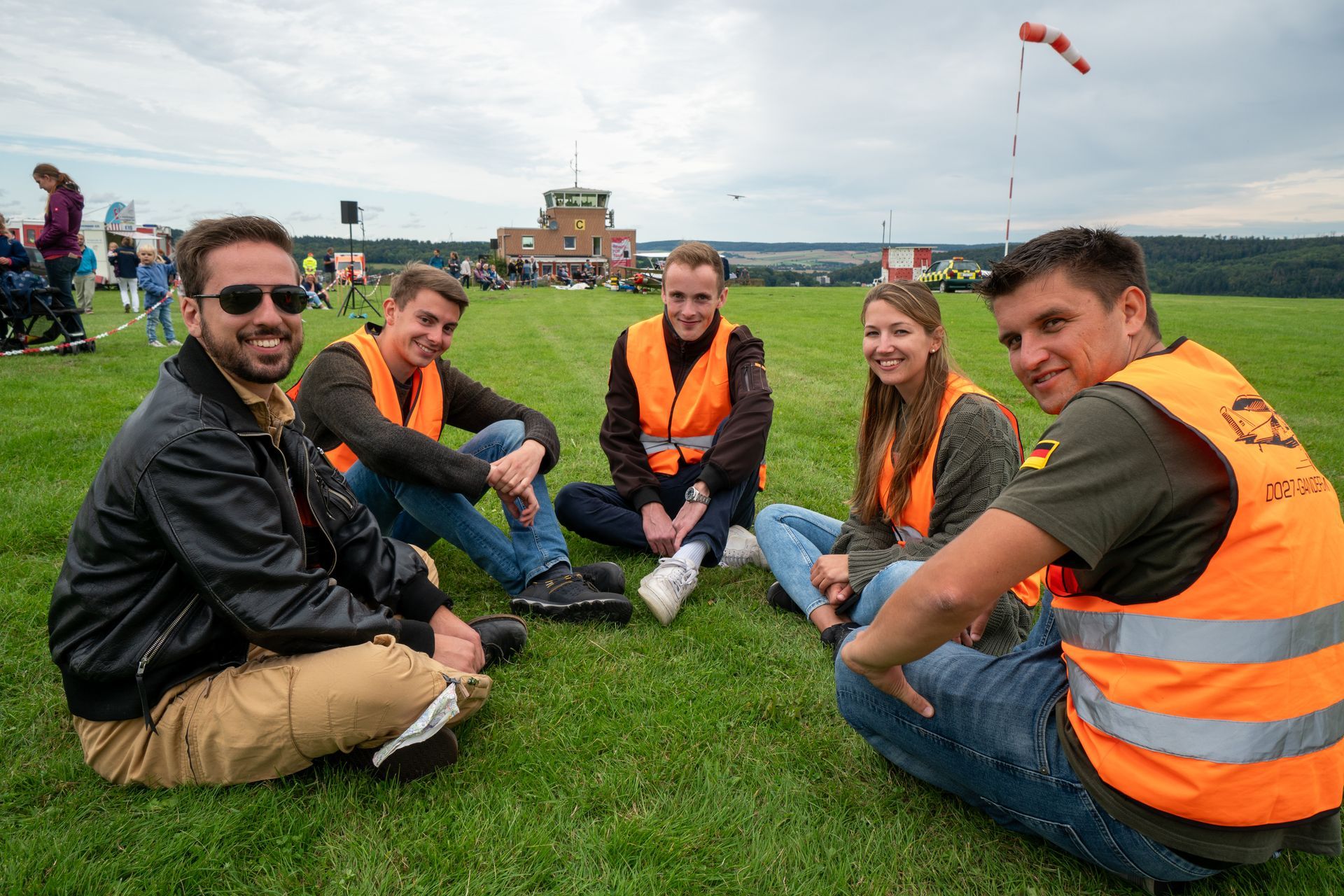 Sechs Personen in orangefarbenen Westen sitzen auf einer Wiese; im Hintergrund ist der Kontrollturm zu sehen.