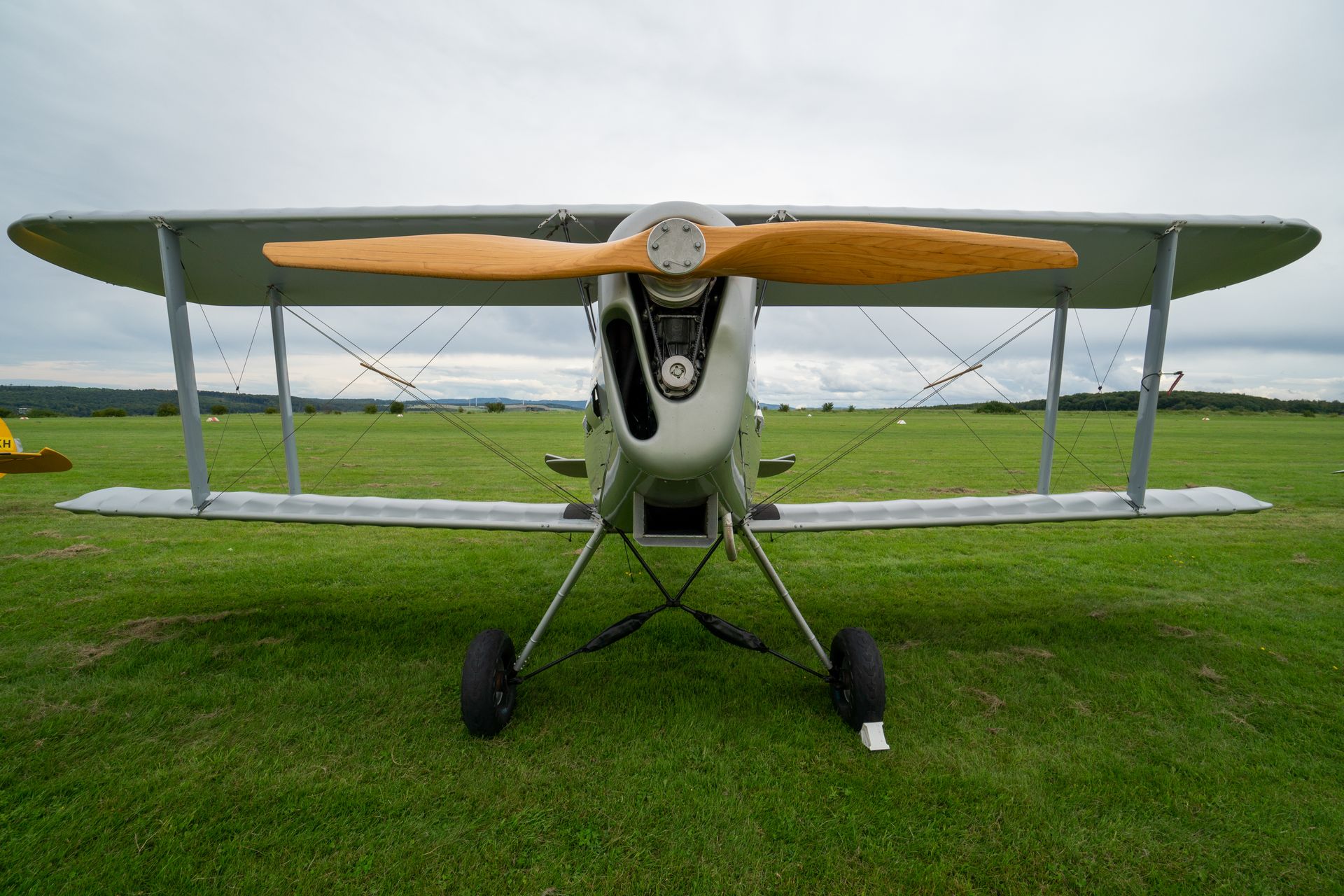 Silbernes Doppeldeckerflugzeug auf einem grasbewachsenen Feld, im Hintergrund ein großer Holzpropeller und ein bewölkter Himmel.