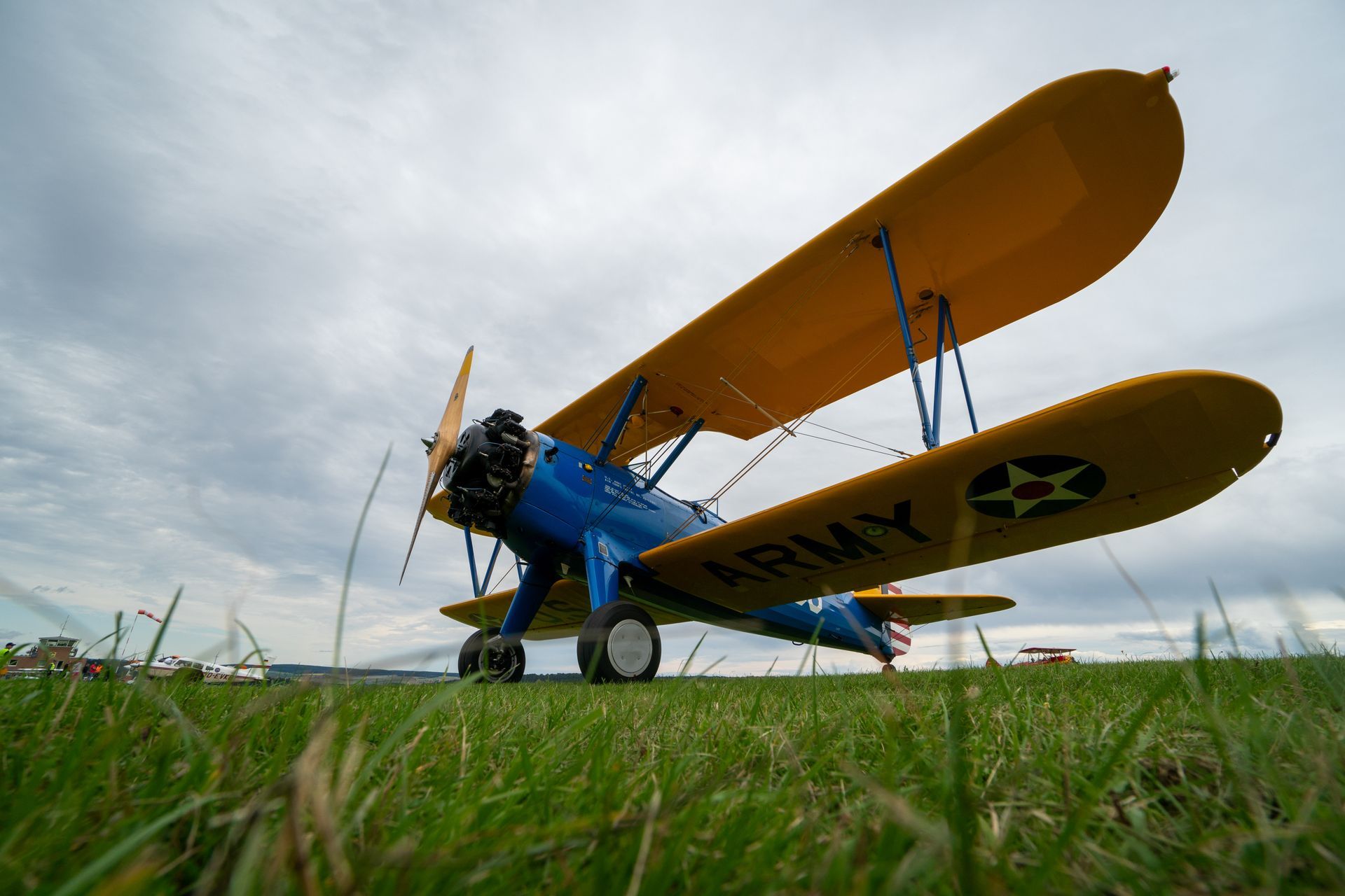 Blau-gelbes Doppeldeckerflugzeug auf grünem Gras vor bewölktem Himmel; an der Seite ist „ARMY“ aufgemalt.