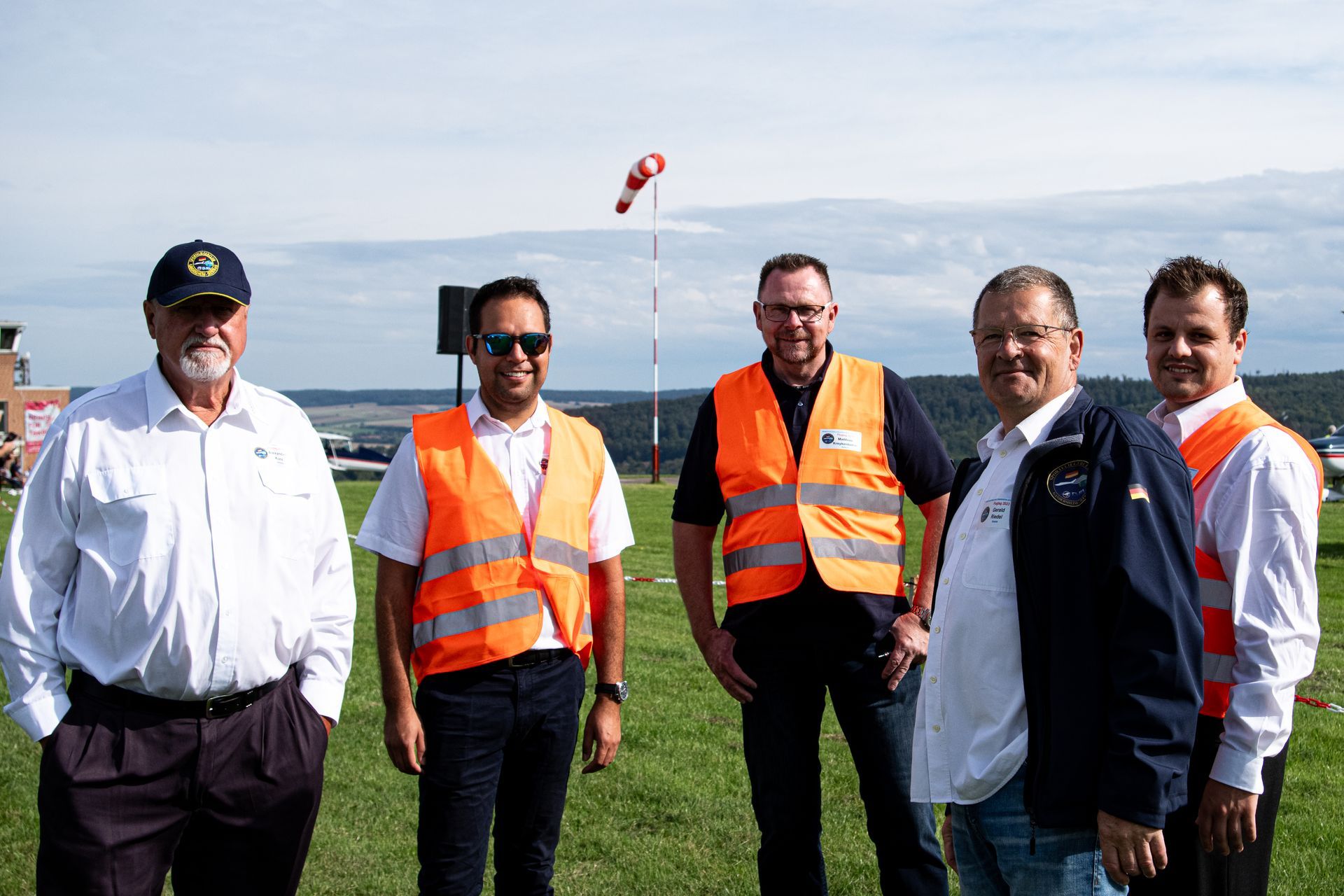 Fünf Männer in leuchtenden Westen posieren auf einem grasbewachsenen Feld; im Hintergrund ist ein Windsack zu sehen.
