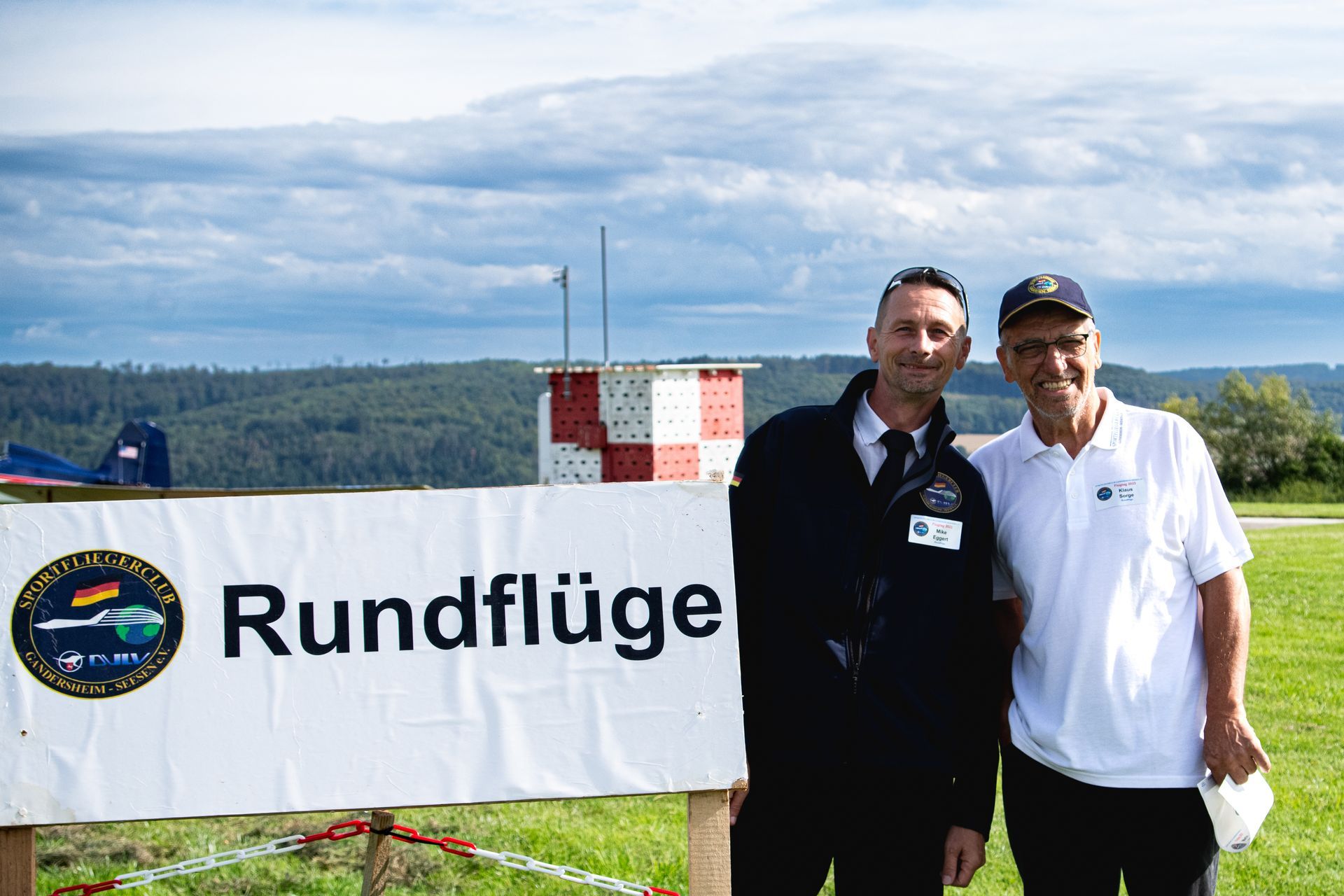 Zwei Männer stehen neben einem Banner mit der Aufschrift „Rundflüge“ auf einem Flugfeld; im Hintergrund sind ein Flugzeug und ein Tower zu sehen.