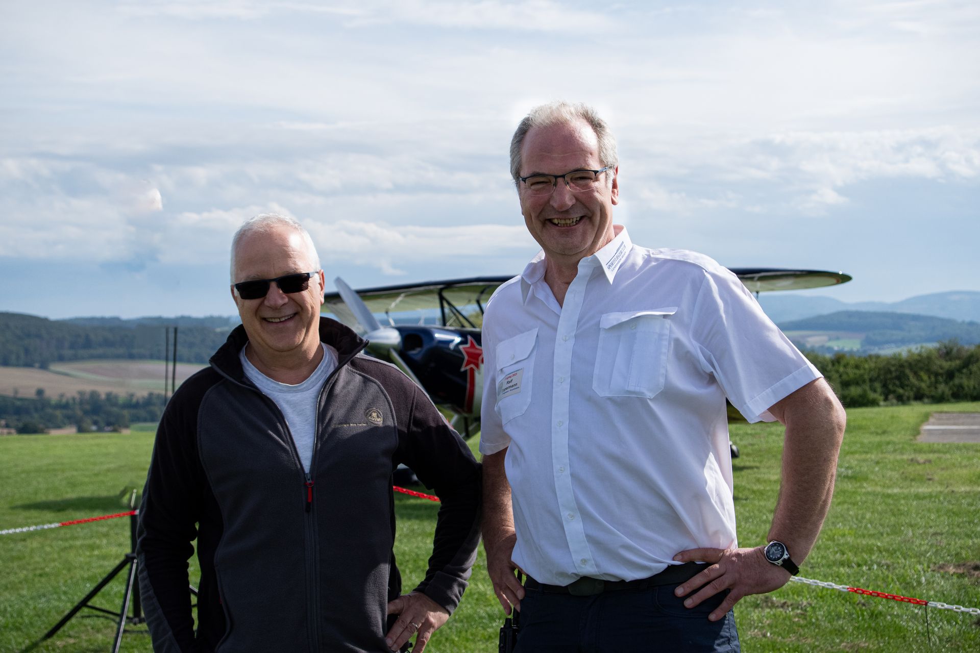Zwei Männer stehen vor einem kleinen Flugzeug auf einem grasbewachsenen Feld; sonniger Tag. Einer der Männer trägt eine Sonnenbrille.