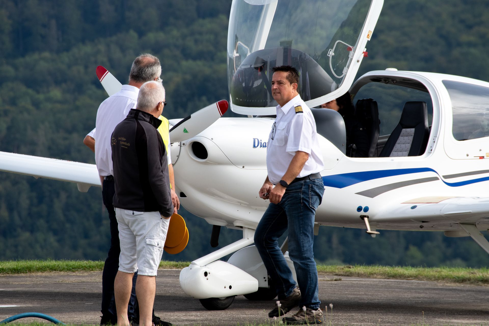 Ein Mann in Pilotenuniform unterhält sich mit zwei Personen in der Nähe eines kleinen Flugzeugs auf dem Rollfeld.