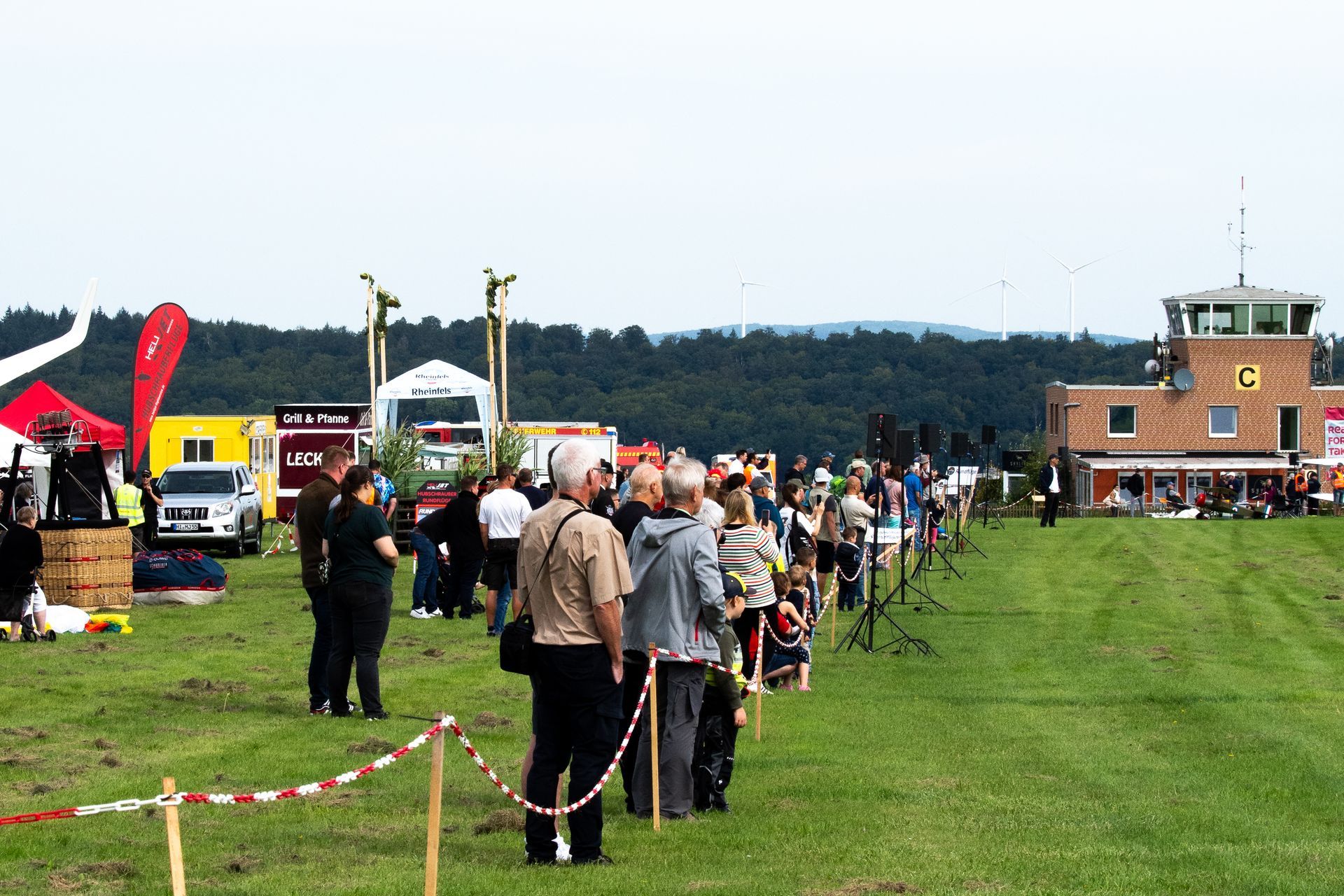 Menschen stehen unter bewölktem Himmel auf einer Rasenfläche in der Nähe eines Kontrollturms und Festivalzelten.