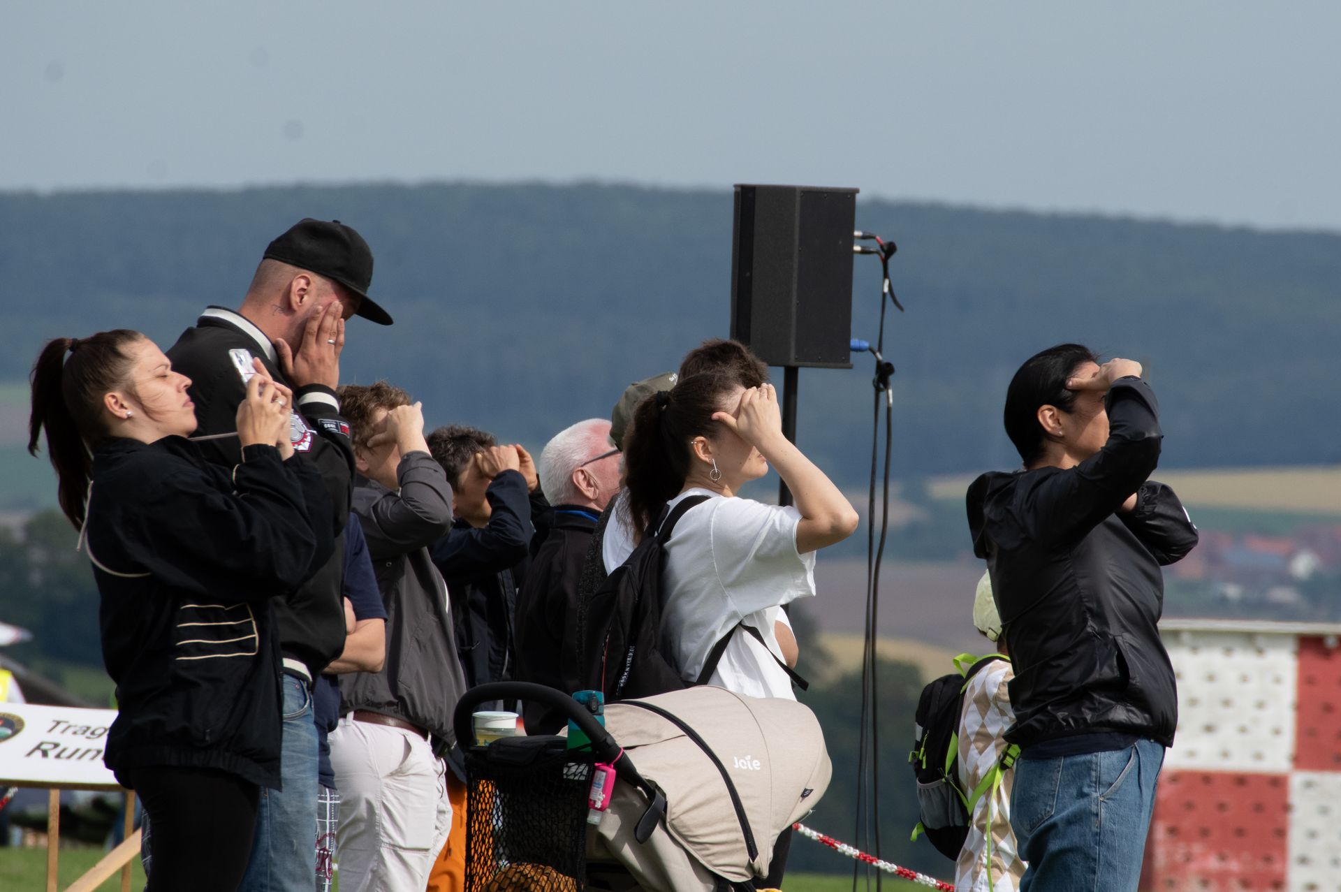 Eine Menschenmenge blickt nach oben, einige halten sich die Hände vor die Augen, bei einer Freiluftveranstaltung mit einem Redner.
