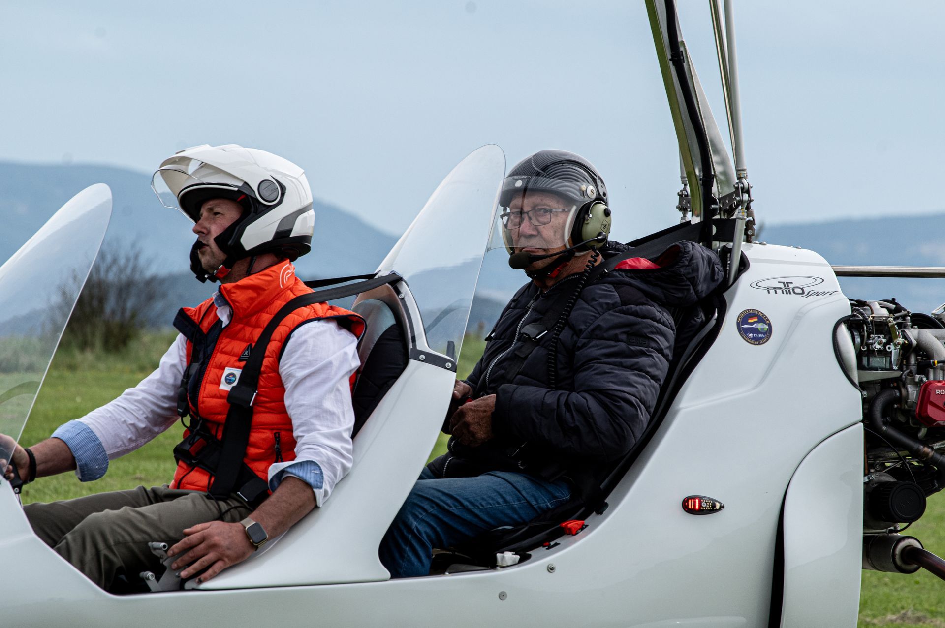 Zwei Männer in einem weißen Tragschrauber, einer am Steuer, beide mit Helmen und Schutzwesten. Grünes Feld und Himmel im Hintergrund.