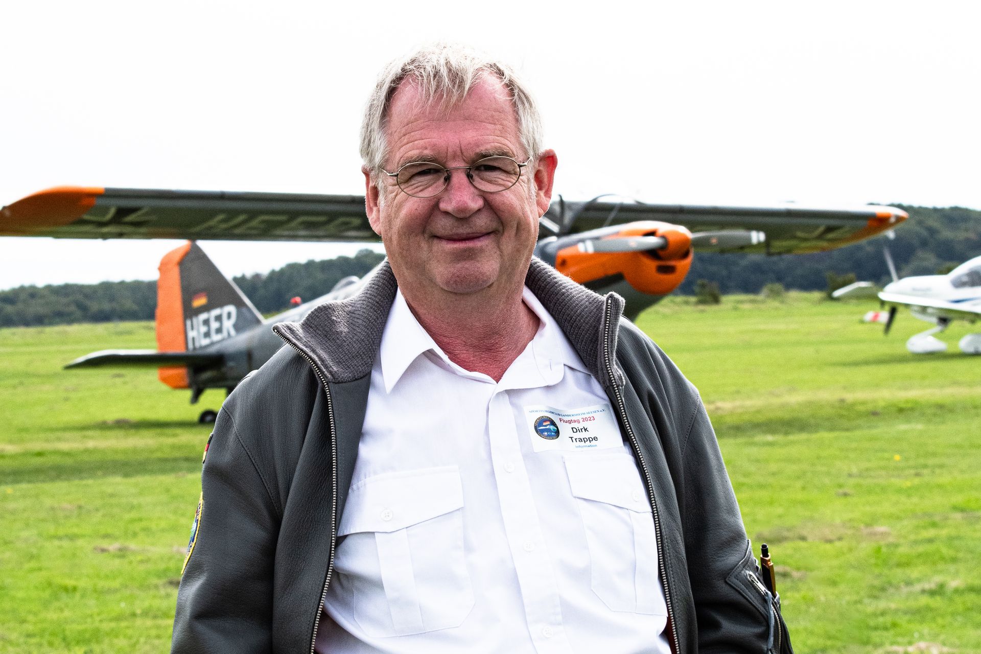 Ein Mann steht vor einem kleinen Flugzeug auf einem grasbewachsenen Feld; er trägt eine Brille, eine Jacke mit Reißverschluss und lächelt.