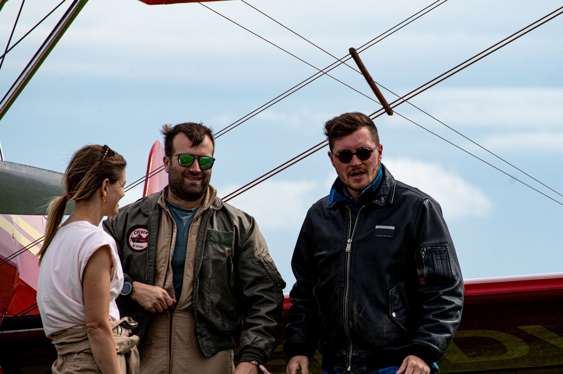 Drei Personen stehen vor einem Doppeldecker; zwei Männer mit Sonnenbrillen und Jacken, eine Frau mit einem hellen Oberteil.