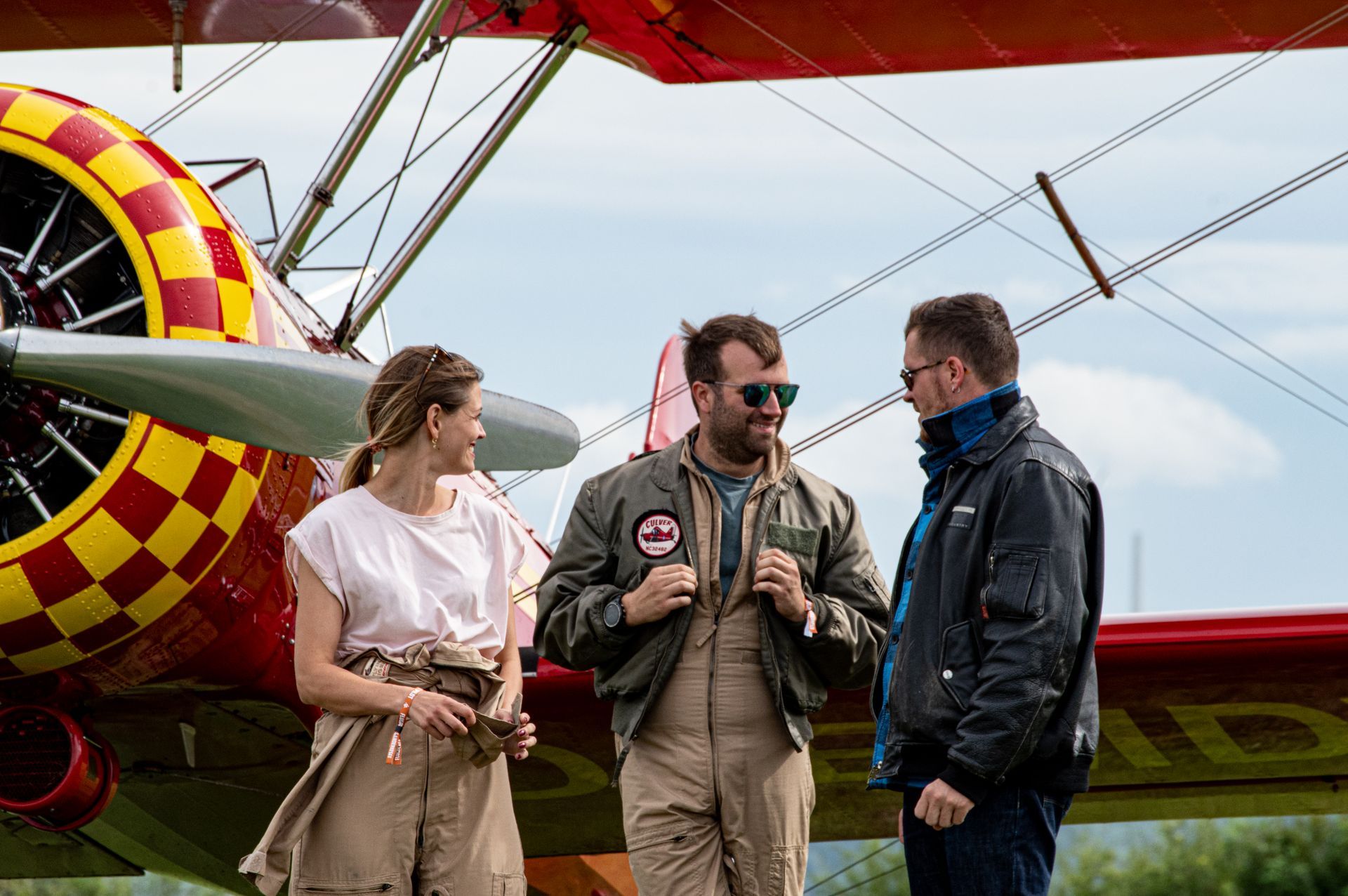 Drei Personen stehen neben einem Doppeldecker mit rot-gelb karierten Tragflächen. Sie unterhalten sich draußen.