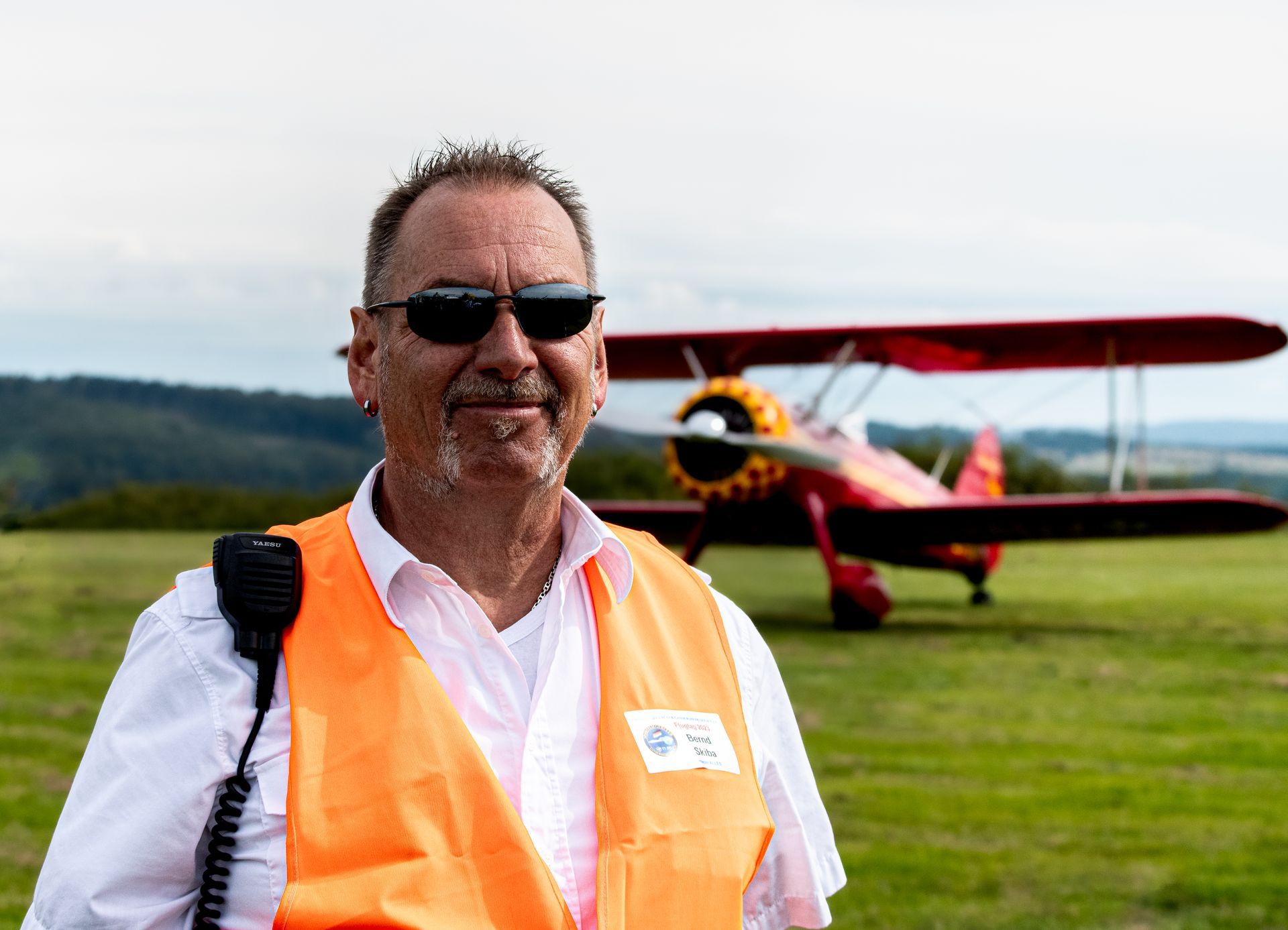 Ein Mann in orangefarbener Weste und Sonnenbrille steht vor einem roten Doppeldecker auf einem grasbewachsenen Feld.