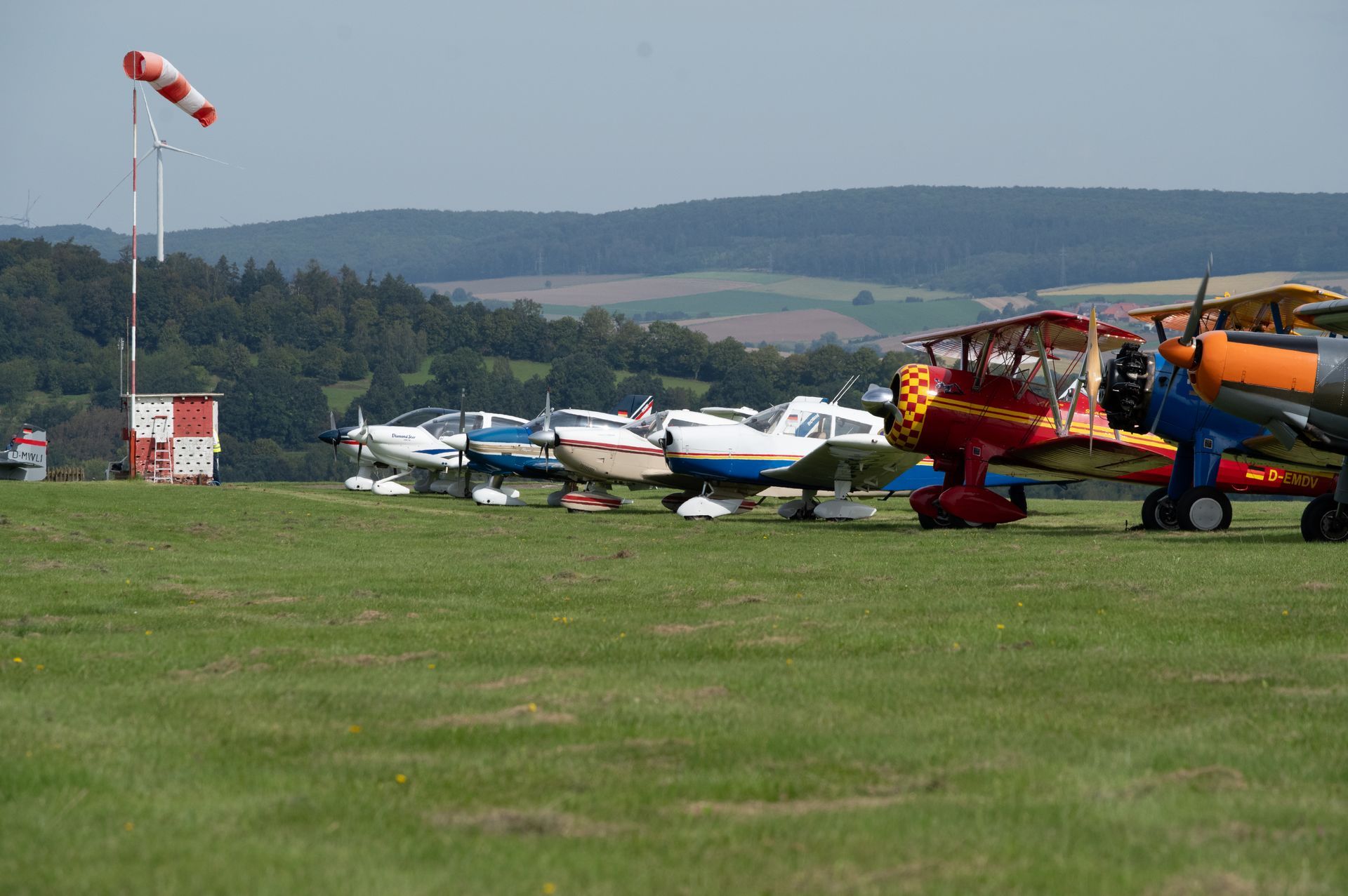 Flugzeuge parken auf einem grasbewachsenen Flugfeld; Windsack und Kontrollturm sind sichtbar.