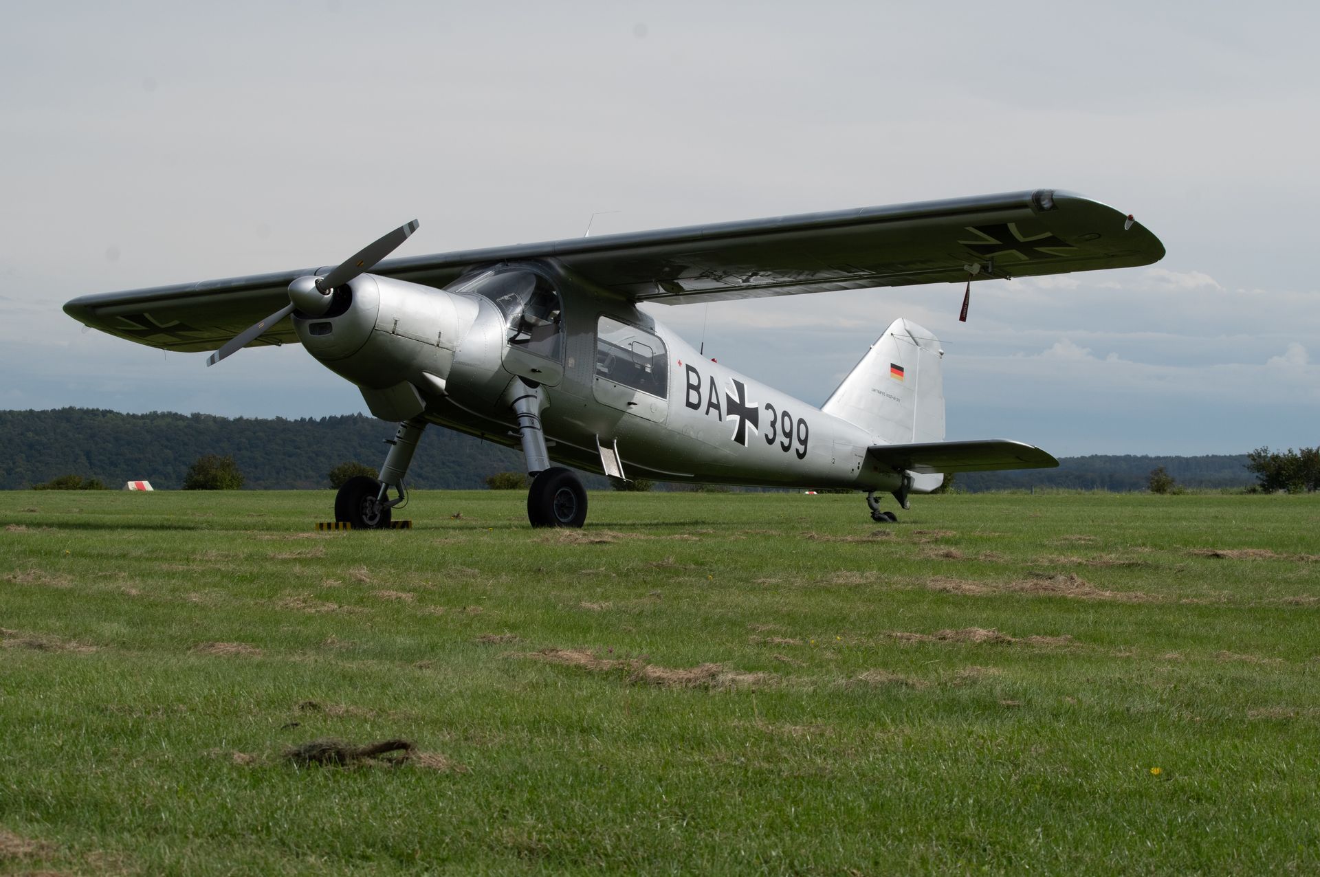 Silbernes, einmotoriges STOL-Flugzeug auf einem Grasfeld. Schwarzer Propeller, deutsche Hoheitsabzeichen.