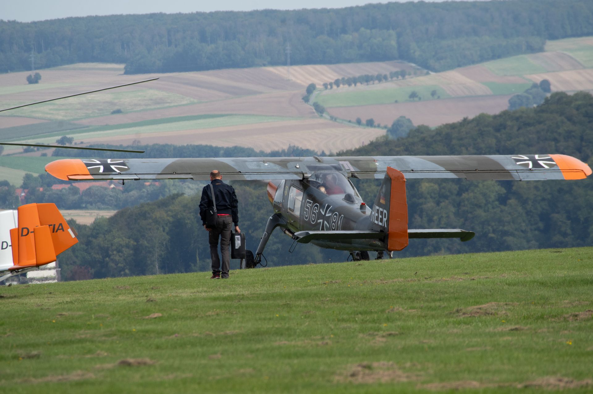 Ein Mann geht auf ein kleines, tarnfarben lackiertes Flugzeug auf einem grasbewachsenen Feld zu; im Hintergrund sind Hügel zu sehen.