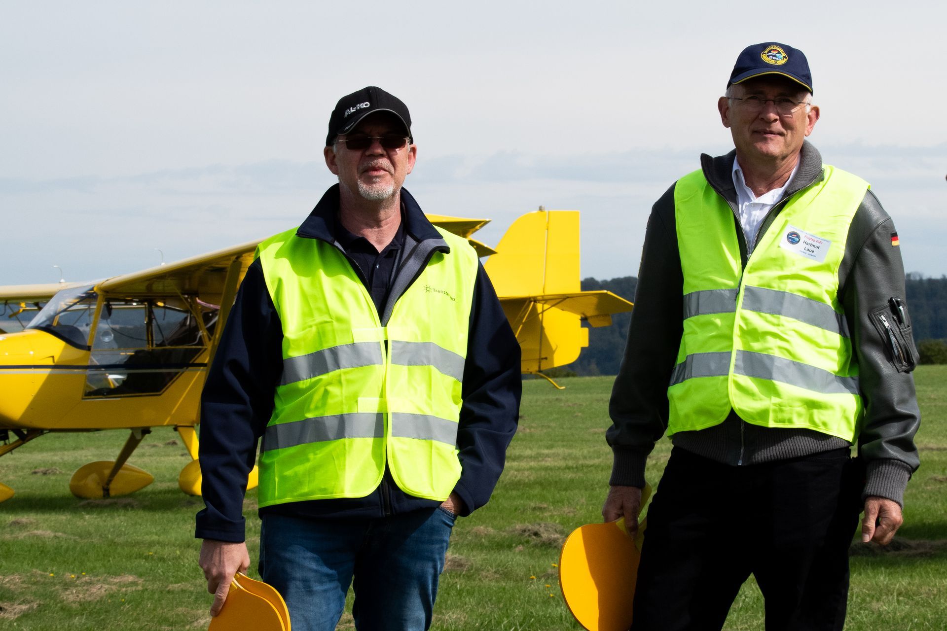 Zwei Männer in Sicherheitswesten stehen neben einem gelben Flugzeug auf einem grasbewachsenen Feld.