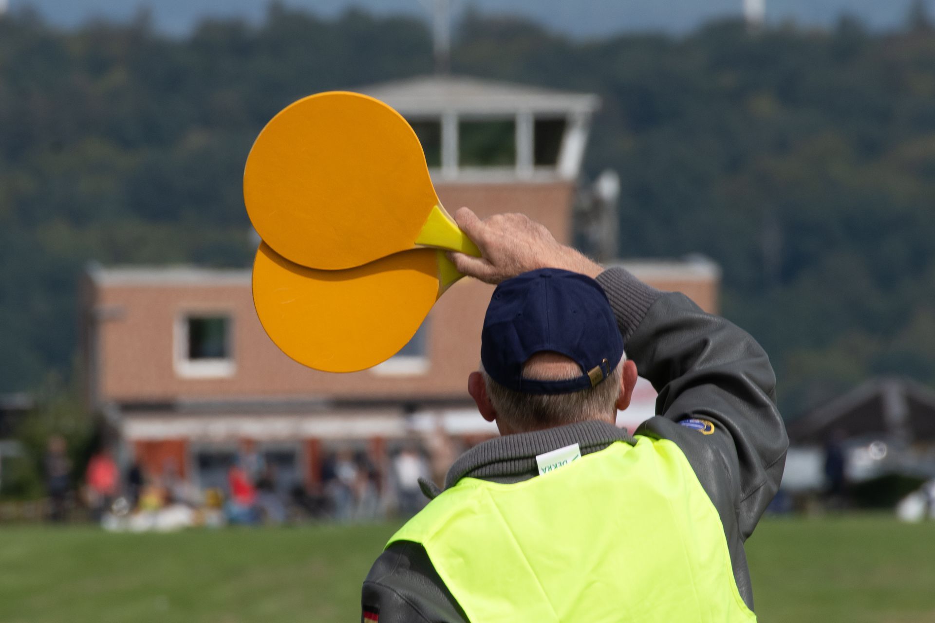 Eine Person in einer gelben Weste, die orangefarbene Paddel hält, gestikuliert in der Nähe eines Kontrollturms auf einem grasbewachsenen Feld.