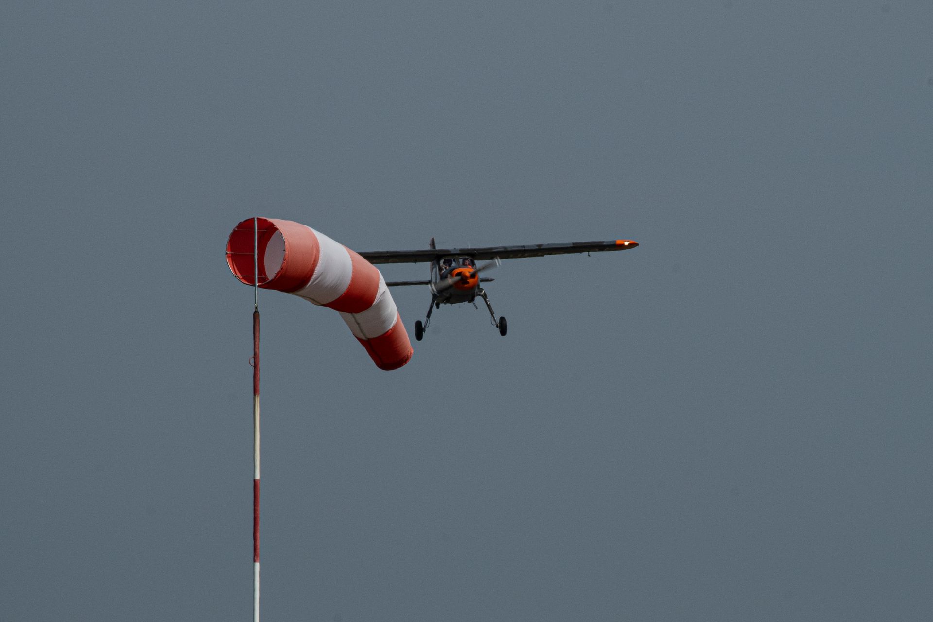 Kleines Flugzeug in der Nähe eines Windsacks; der Windsack ist rot-weiß. Grauer Himmel im Hintergrund.