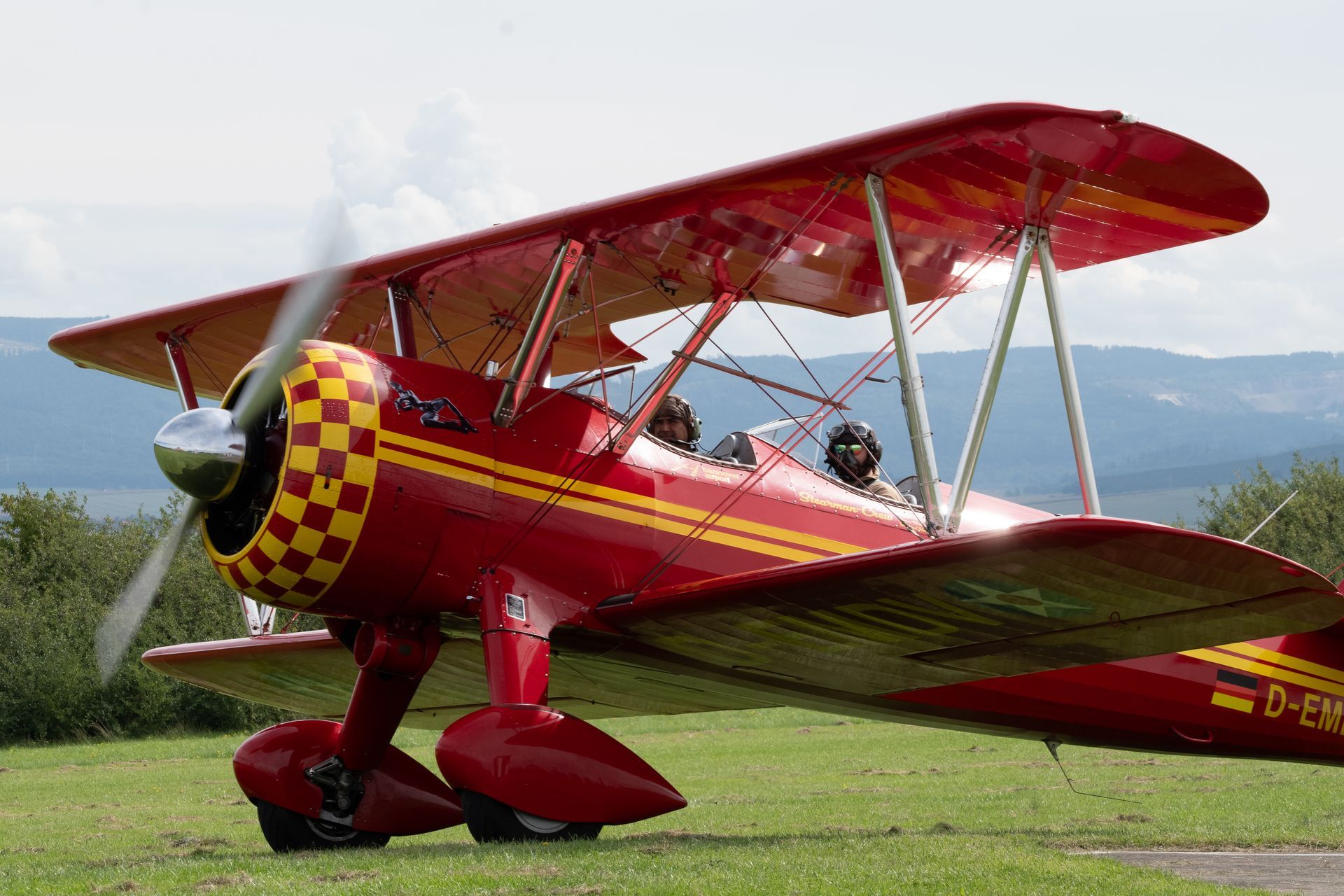 Ein rot-gelbes Doppeldeckerflugzeug auf einem grasbewachsenen Feld, der Propeller dreht sich.