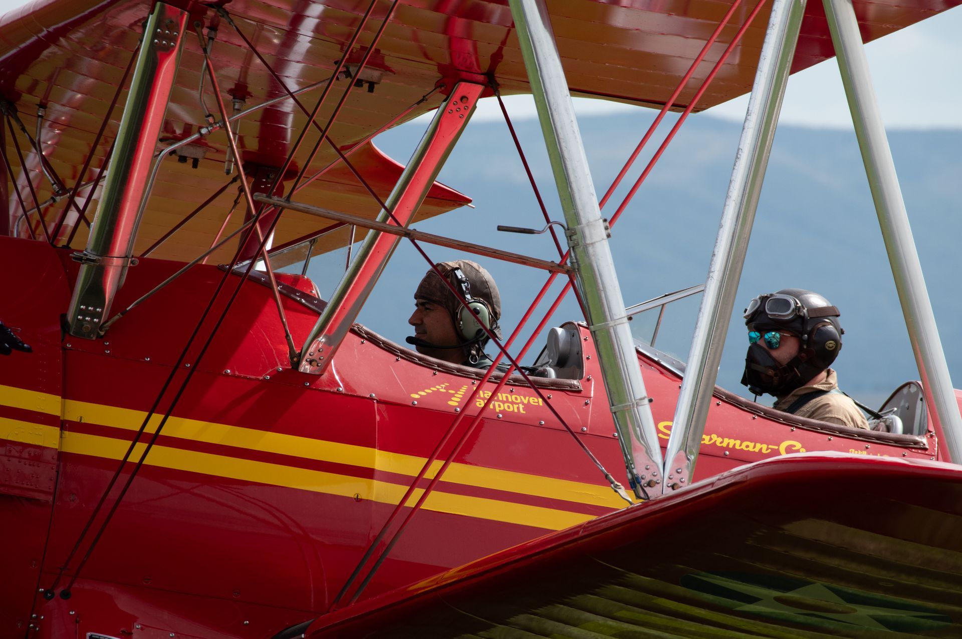 Rotes Doppeldeckerflugzeug mit zwei Piloten in Helmen, Cockpitansicht.