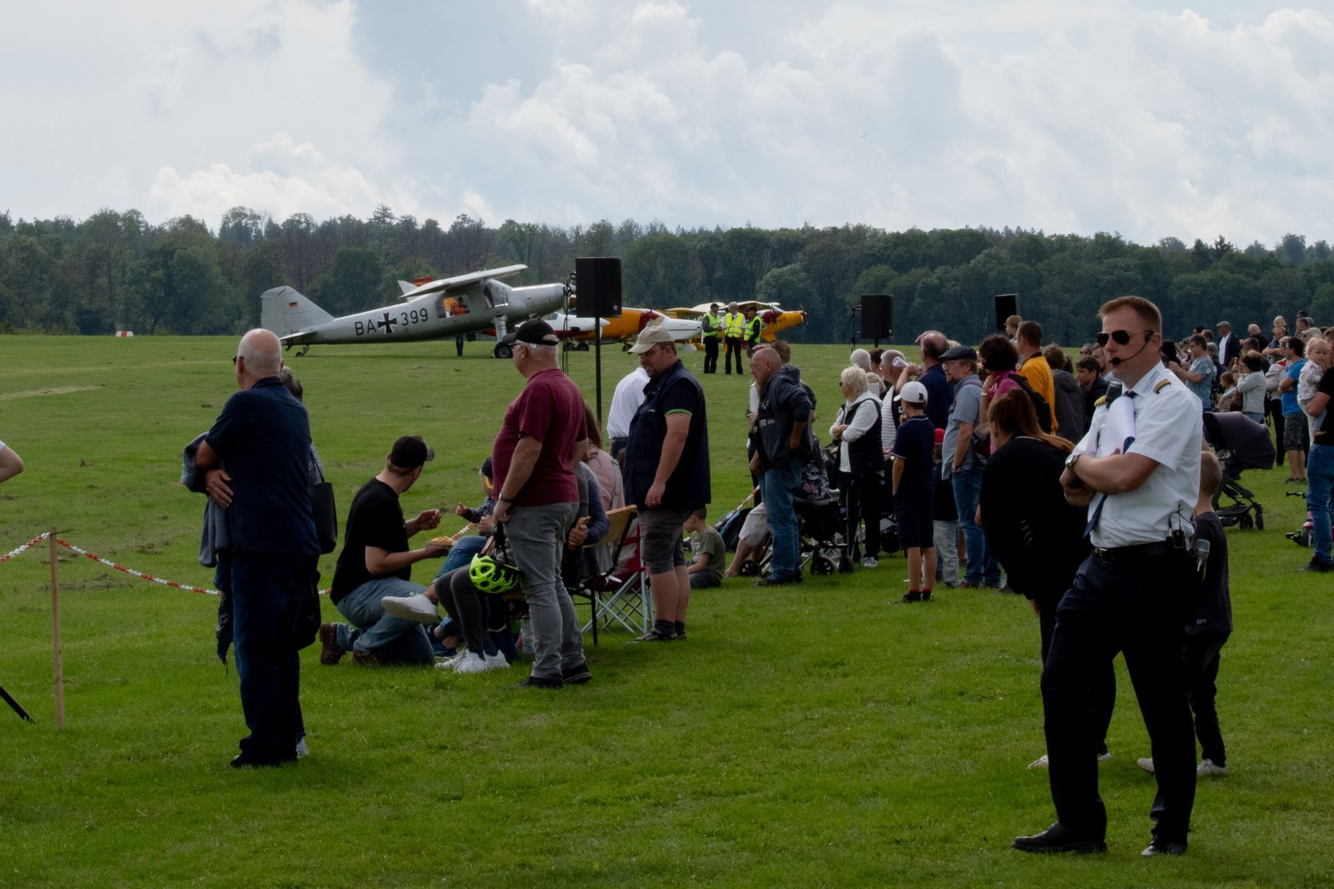 Menschen beobachten ein Flugzeug auf einer Wiese. Einige stehen, andere sitzen. Im Hintergrund sind Bäume zu sehen. Bewölkter Himmel.
