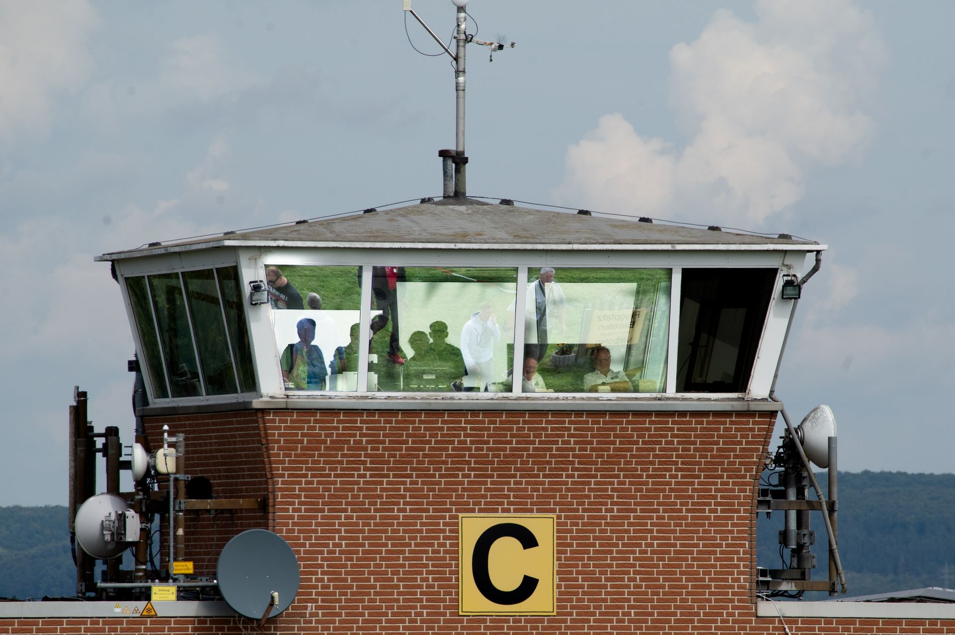 Flugsicherungsturm mit roter Backsteinfassade; durch die Fenster kann man die Personen im Inneren sehen; Satellitenschüssel angebracht.